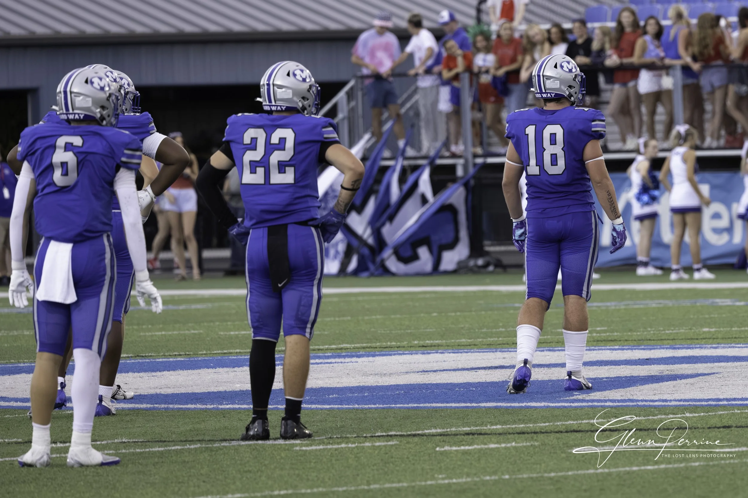 American football players in blue uniforms standing on the field during a game, with spectators watching from the sidelines.