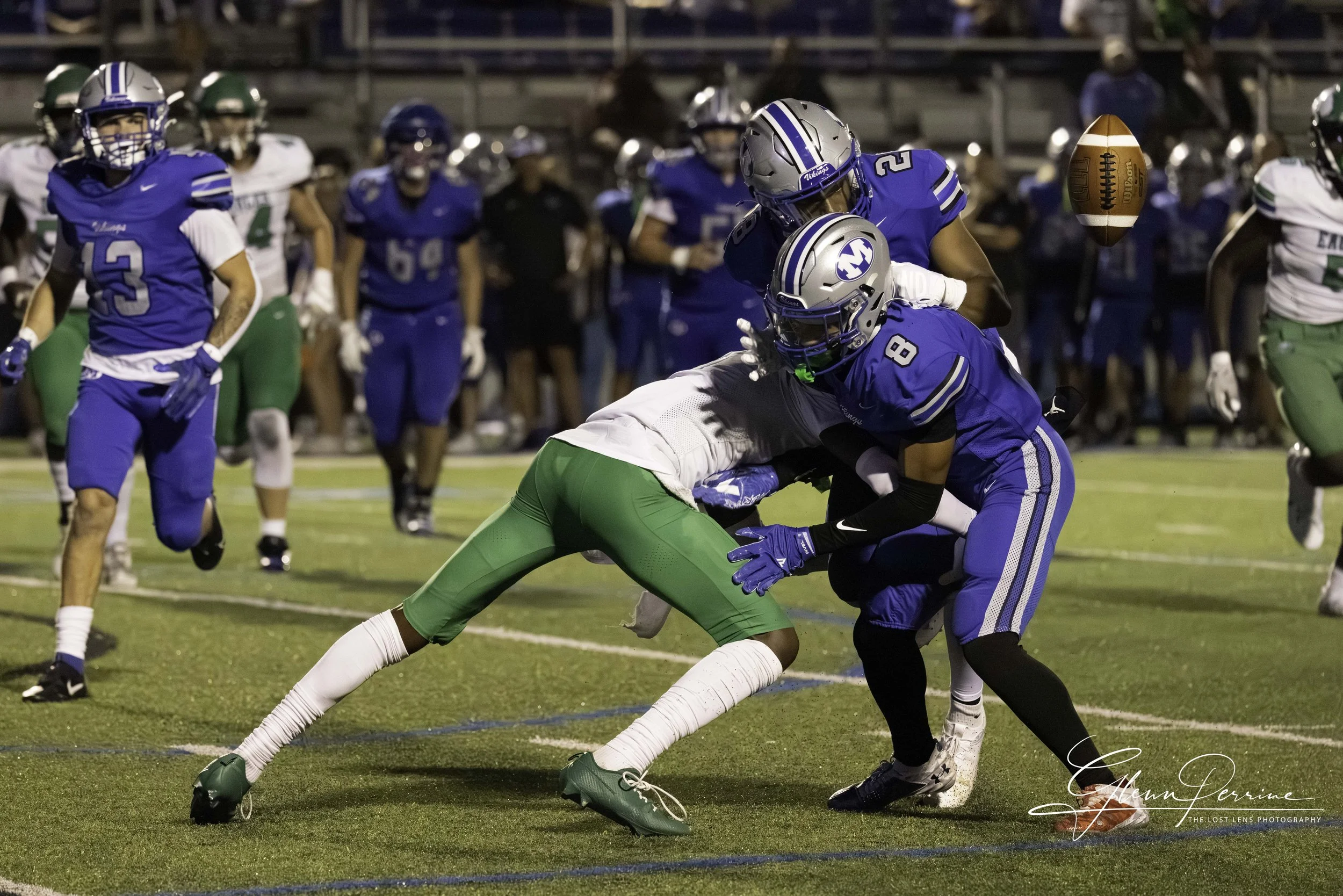Football game with players in blue and green uniforms, two players tackling each other on the field, and others in the background.