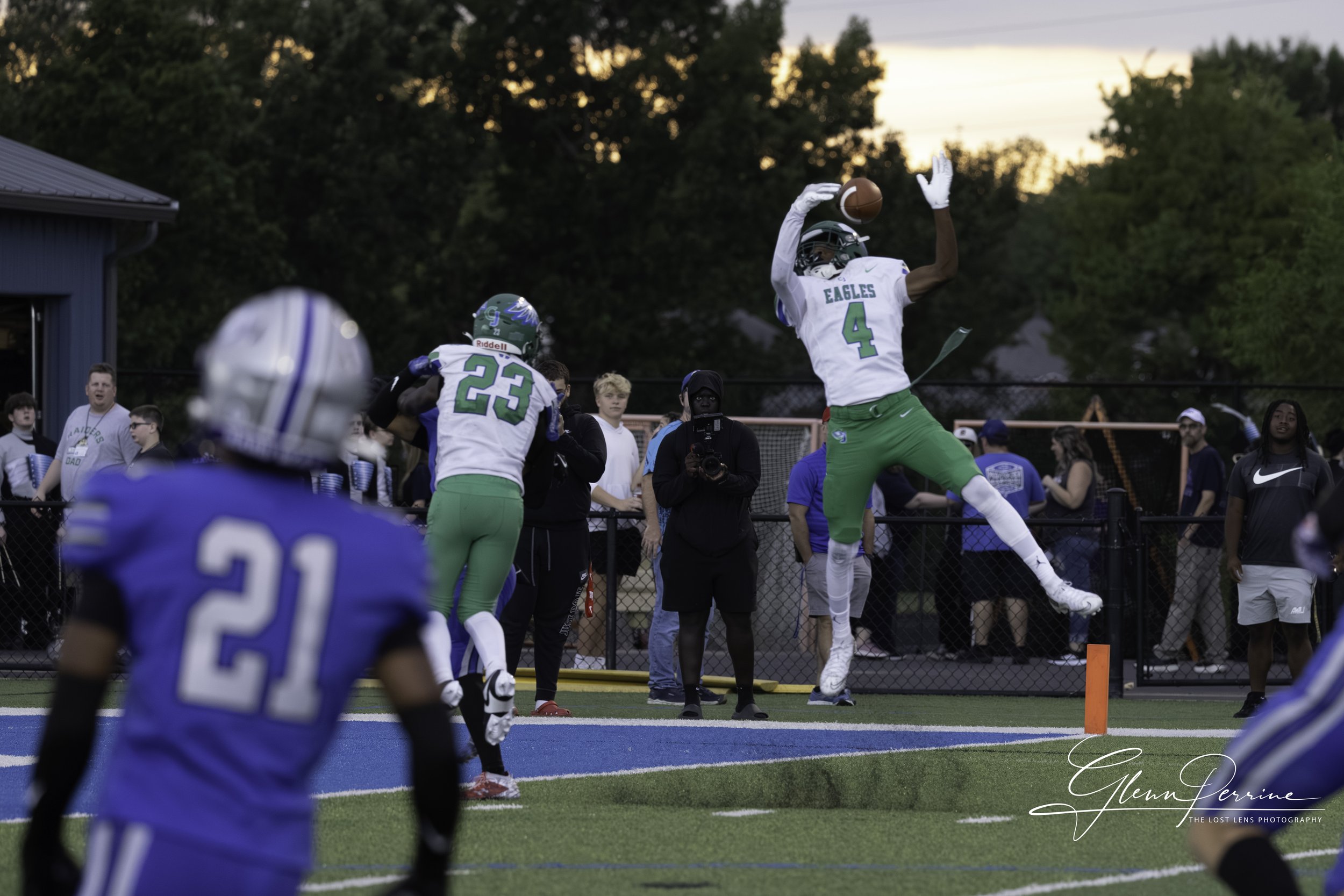 An American football game during sunset with a player catching a touchdown pass at the end zone, surrounded by opposing team players and spectators.