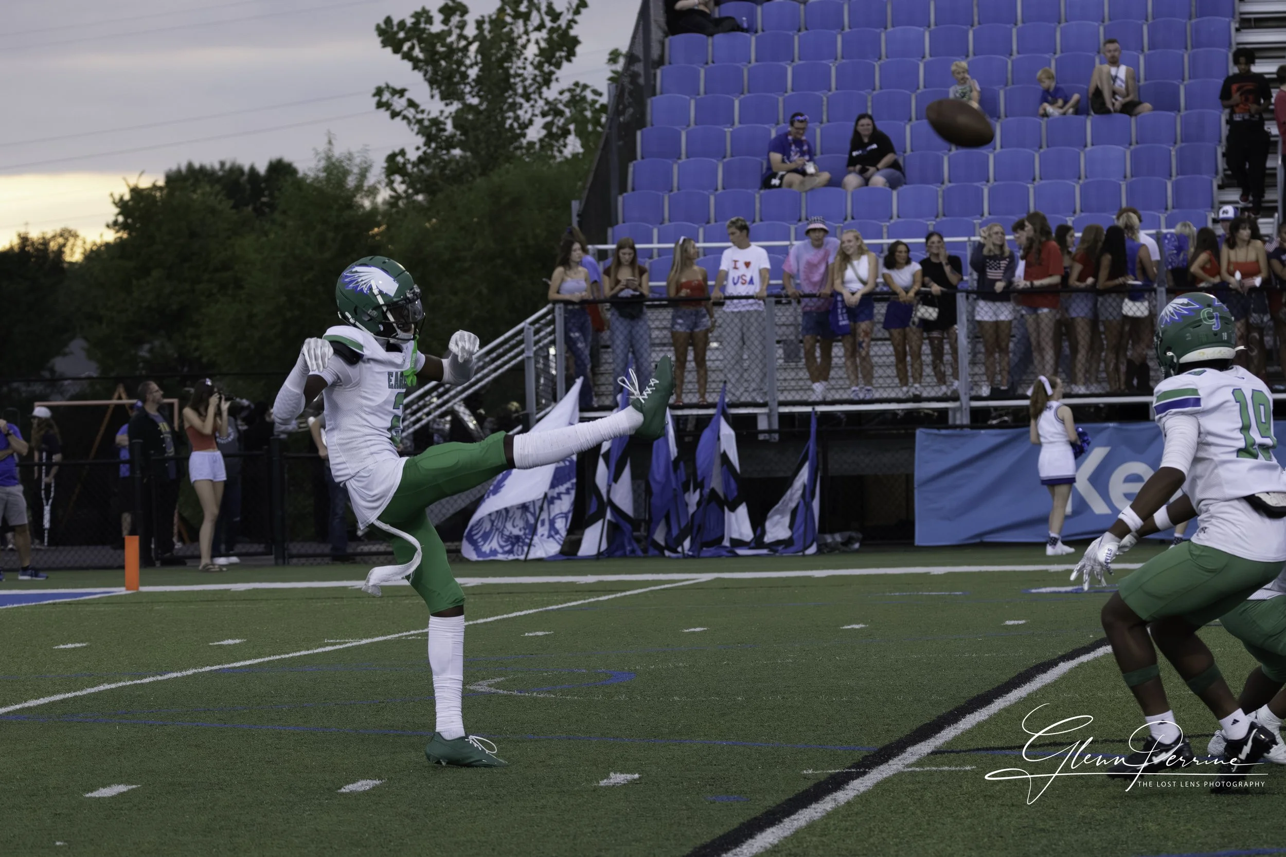 A football game scene with two players in green and white uniforms and helmets, one of them kicking the football, on a field with a crowd of spectators in the background, some sitting in the bleachers and some standing along the sidelines.