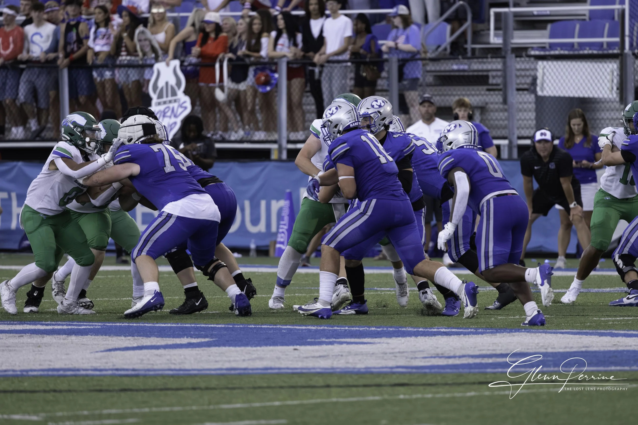 American football game with players in blue and green uniforms engaging at the line of scrimmage, and spectators in the stands in the background.