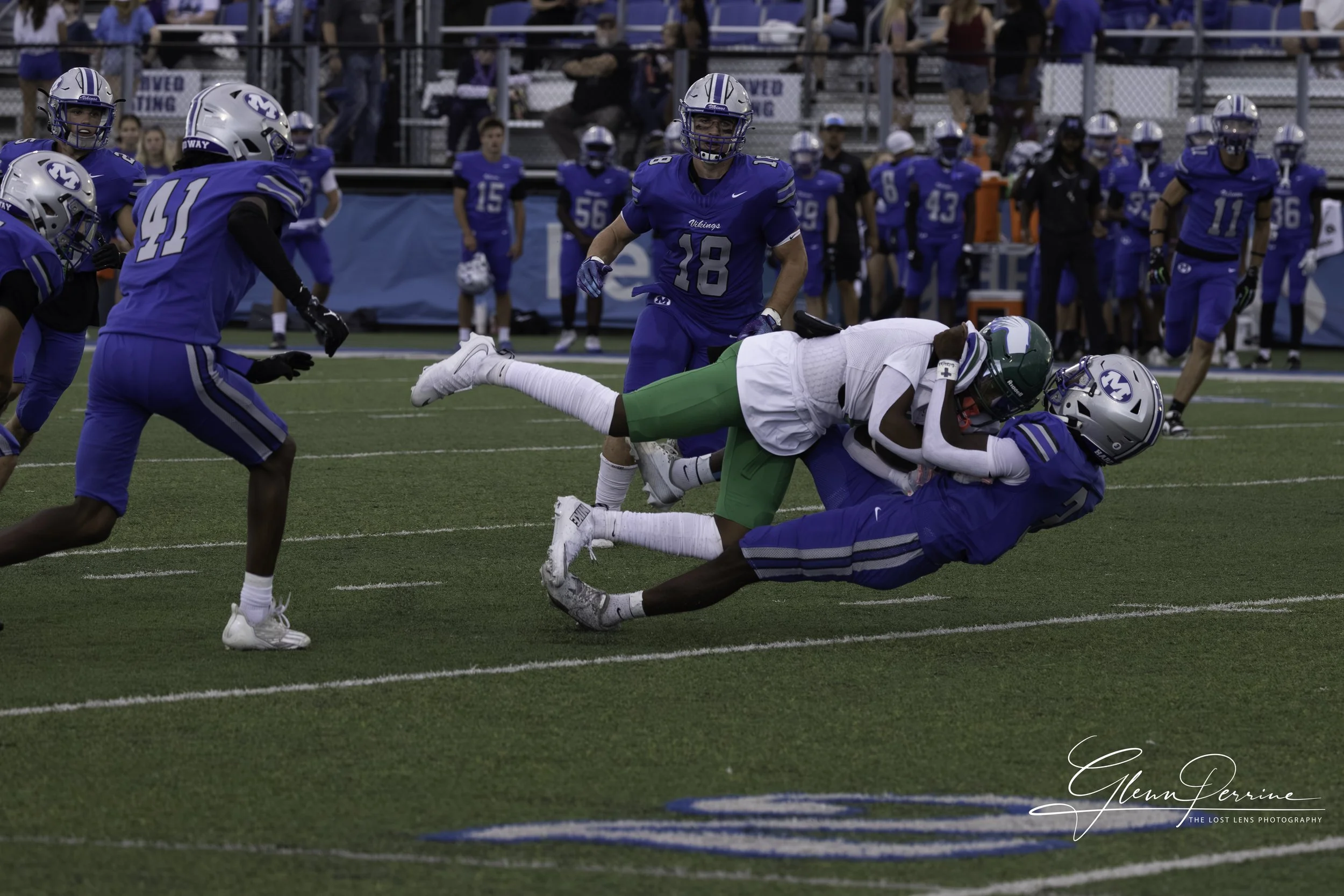 A football game with players in blue and white uniforms. One player is tackled by a player in white and green, while others watch and move on the field.