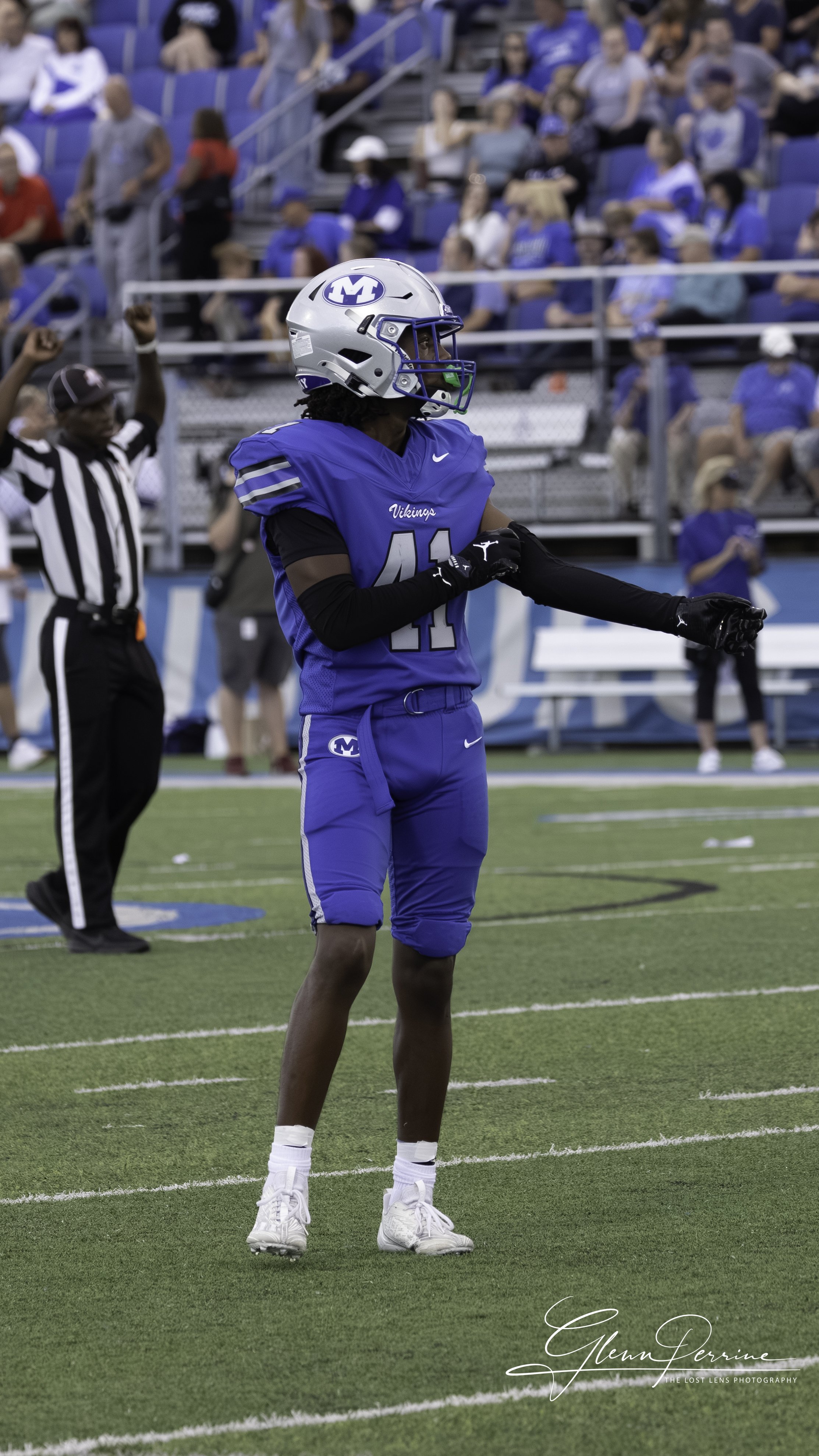 Young football player in a blue uniform and helmet on the field, with spectators in the stands in the background.