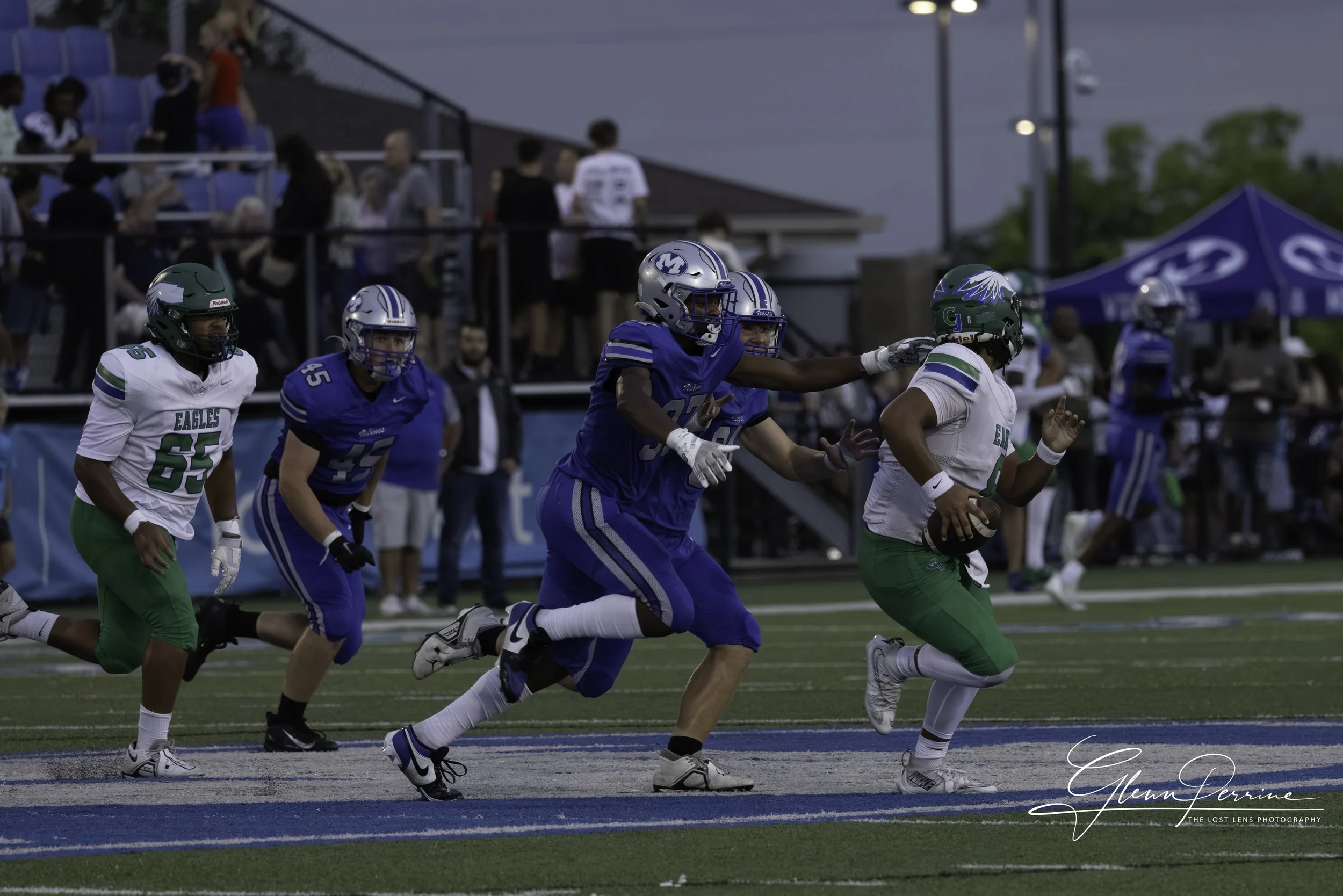 Football players in blue and green uniforms on a field during a game, with spectators watching from the stands.