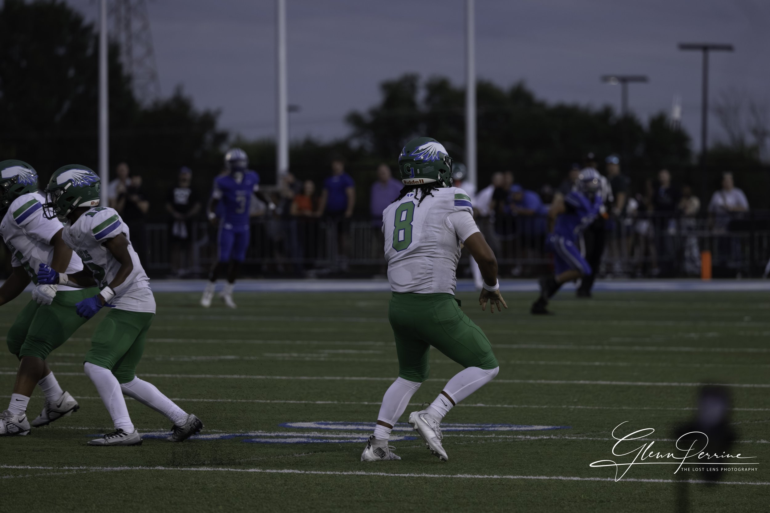Football players on the field during a game, with one player in a white and green uniform preparing to run, with other players and spectators in the background.