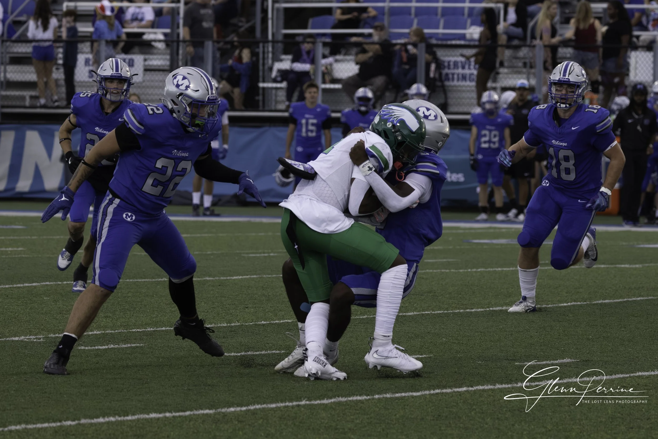 Two football players from the team in white and green are tackled by a player in blue, while other players and spectators are visible in the background.