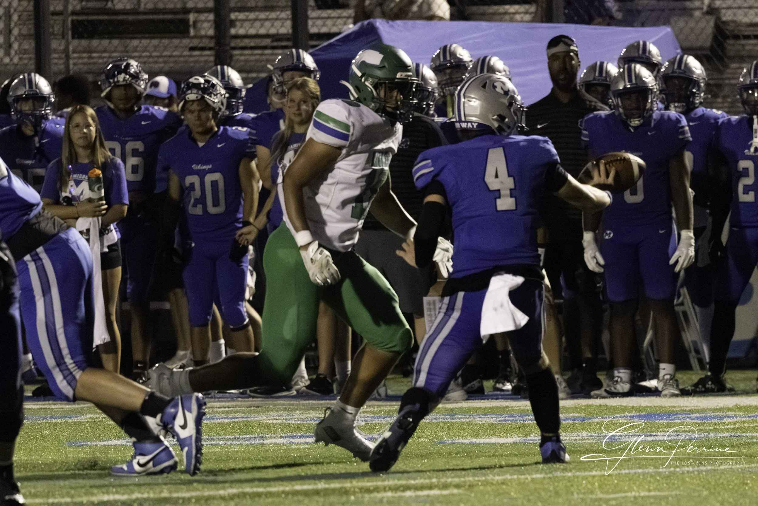 Football game with players in blue and one in green, with a girl holding a flower on the sidelines and other players watching.