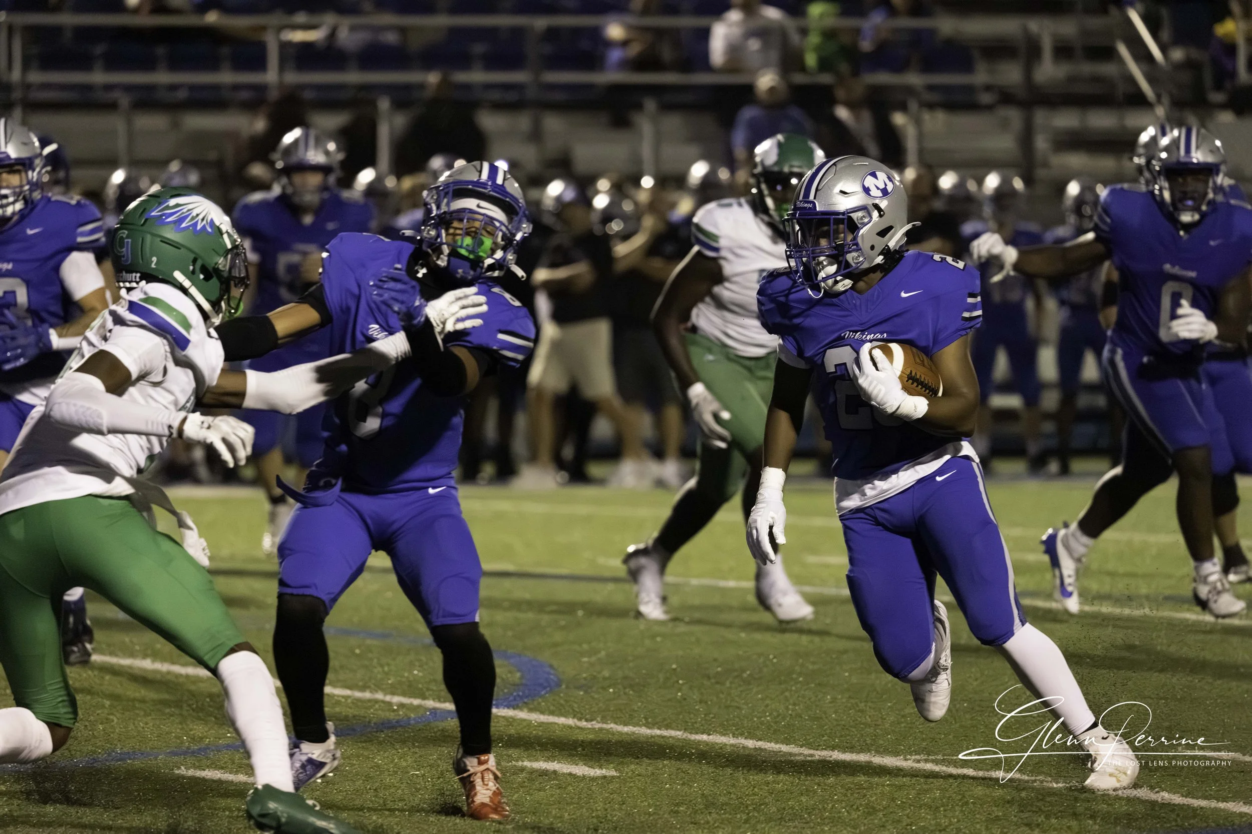 Football game with players in blue and green uniforms on the field, some players blocking and others running with the ball, under stadium lights at night.
