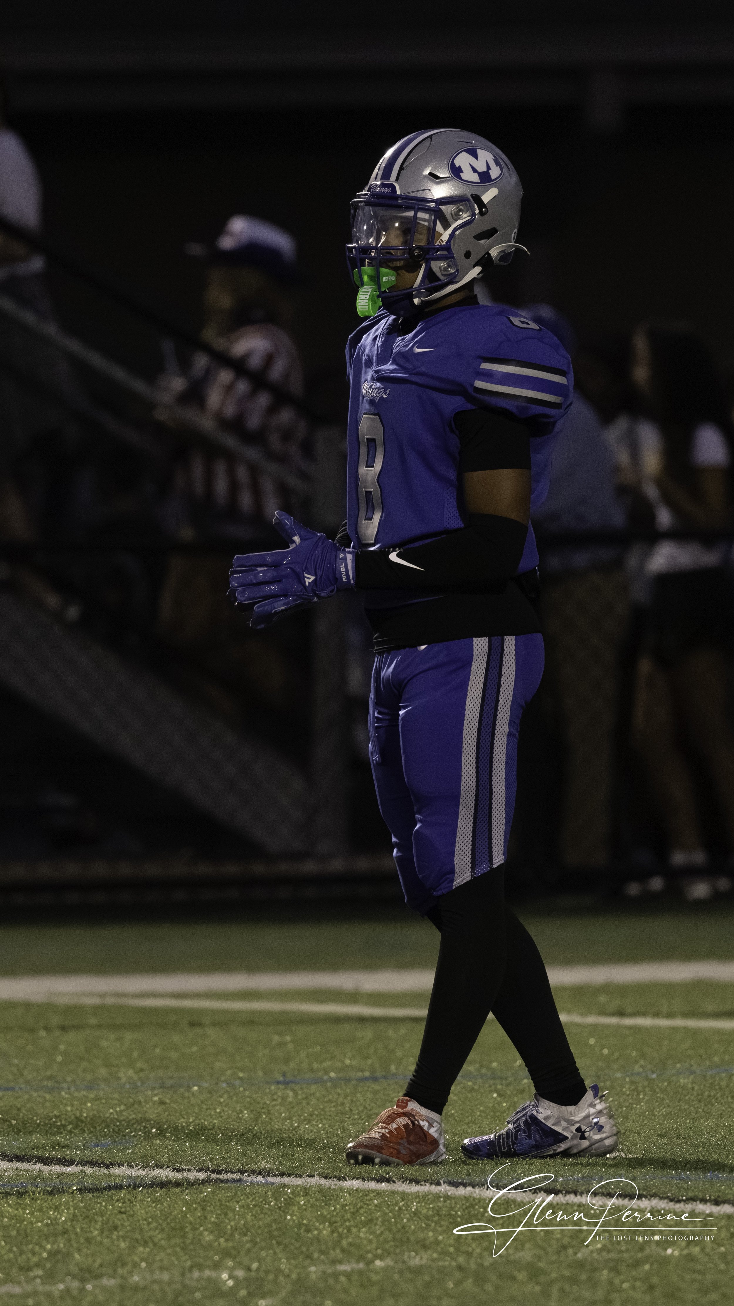 A football player in a blue uniform standing on the field during a game, wearing a helmet with a face mask, gloves, and cleats, with spectators in the background.