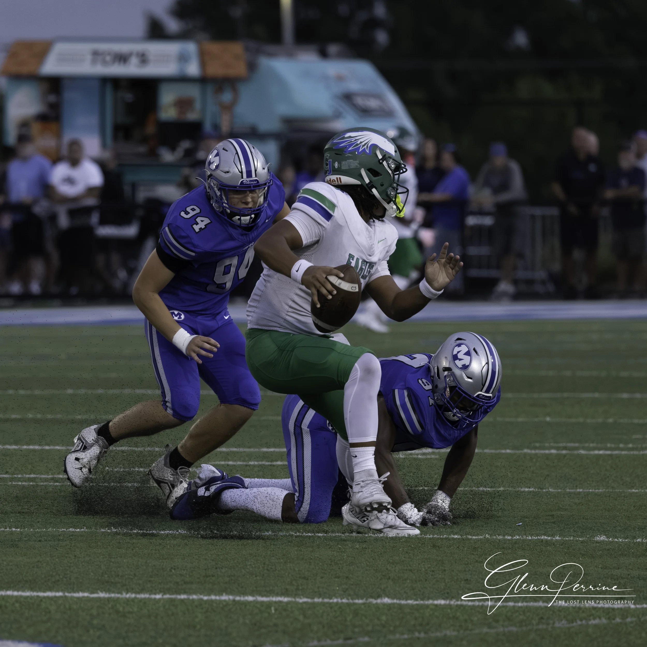 A football player in a white and green uniform running with the ball, being tackled by two players in blue uniforms on a football field, with spectators in the background.