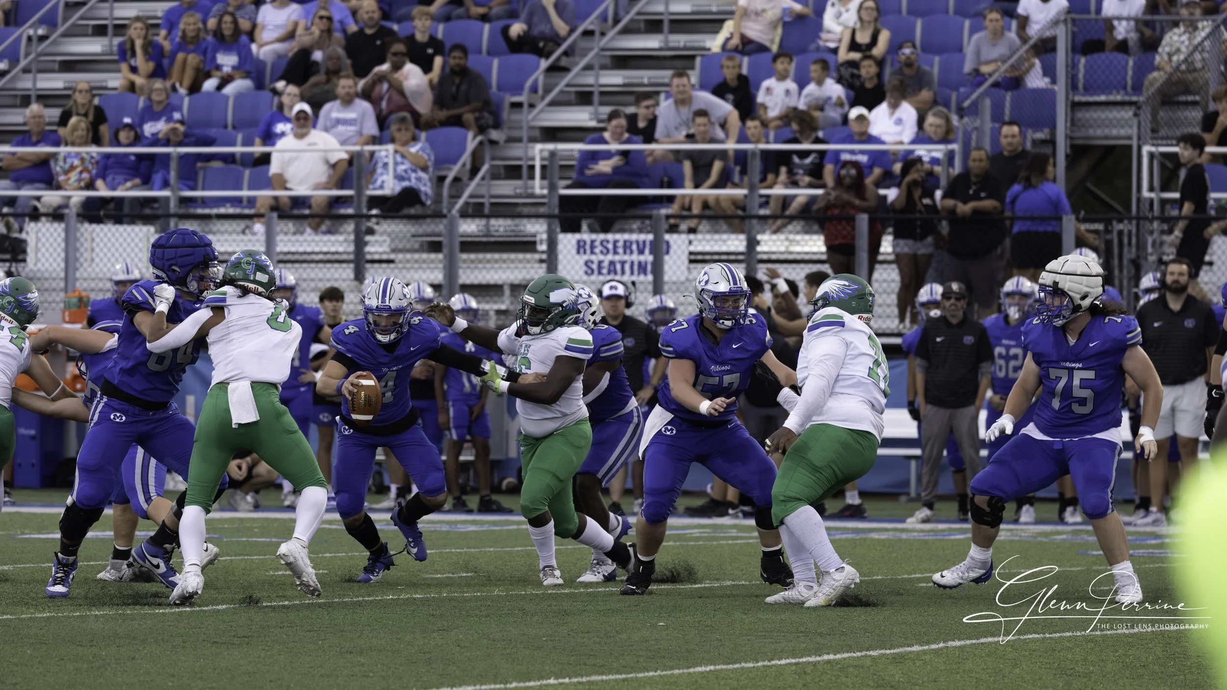 A football game with players in blue and white uniforms on the field, with spectators sitting in the stands behind a fence, some in reserved seating area, watching the game.