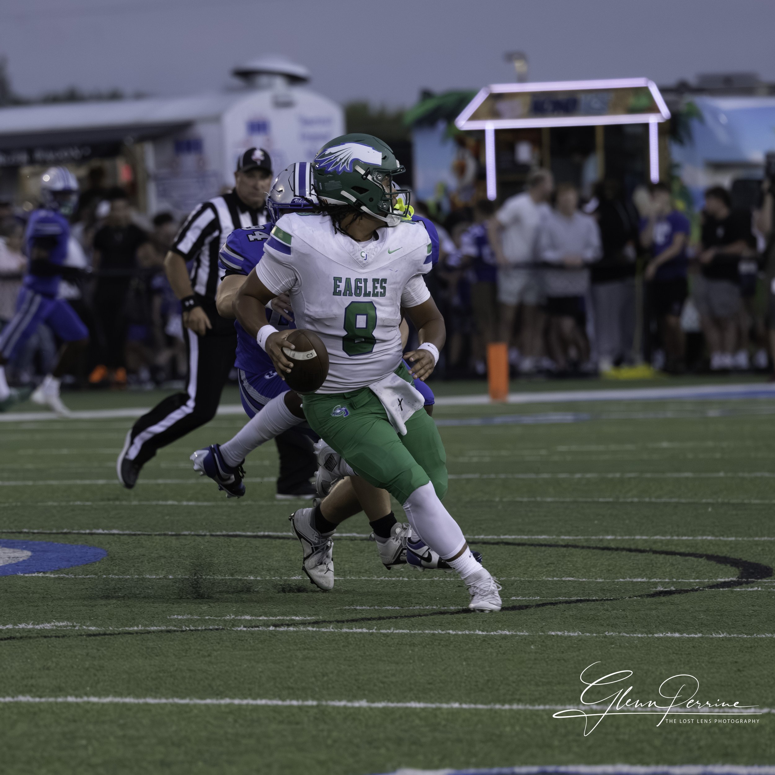 An American football player in a white and green uniform with the number 8, running with the football while a blue defender attempts to tackle him during a game on a football field.