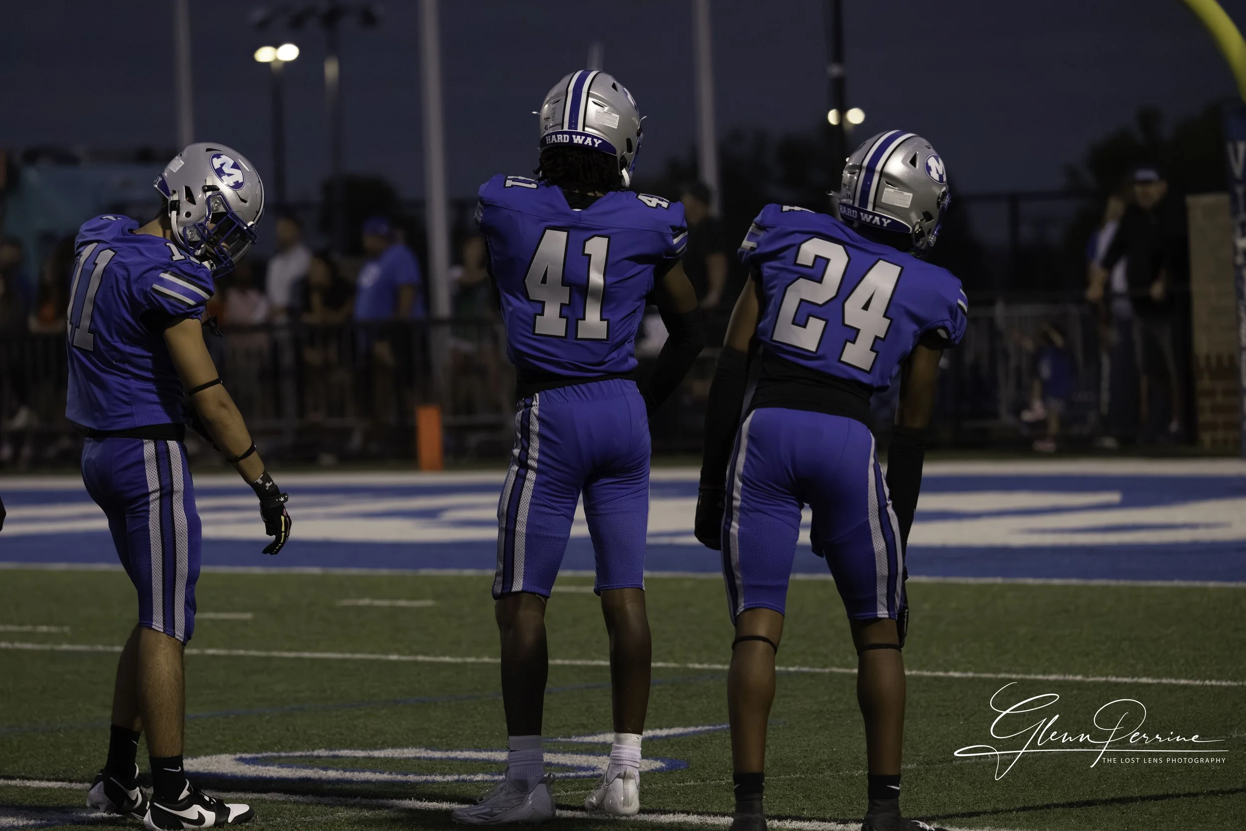 Three football players in blue uniforms standing on a football field during a game at dusk.