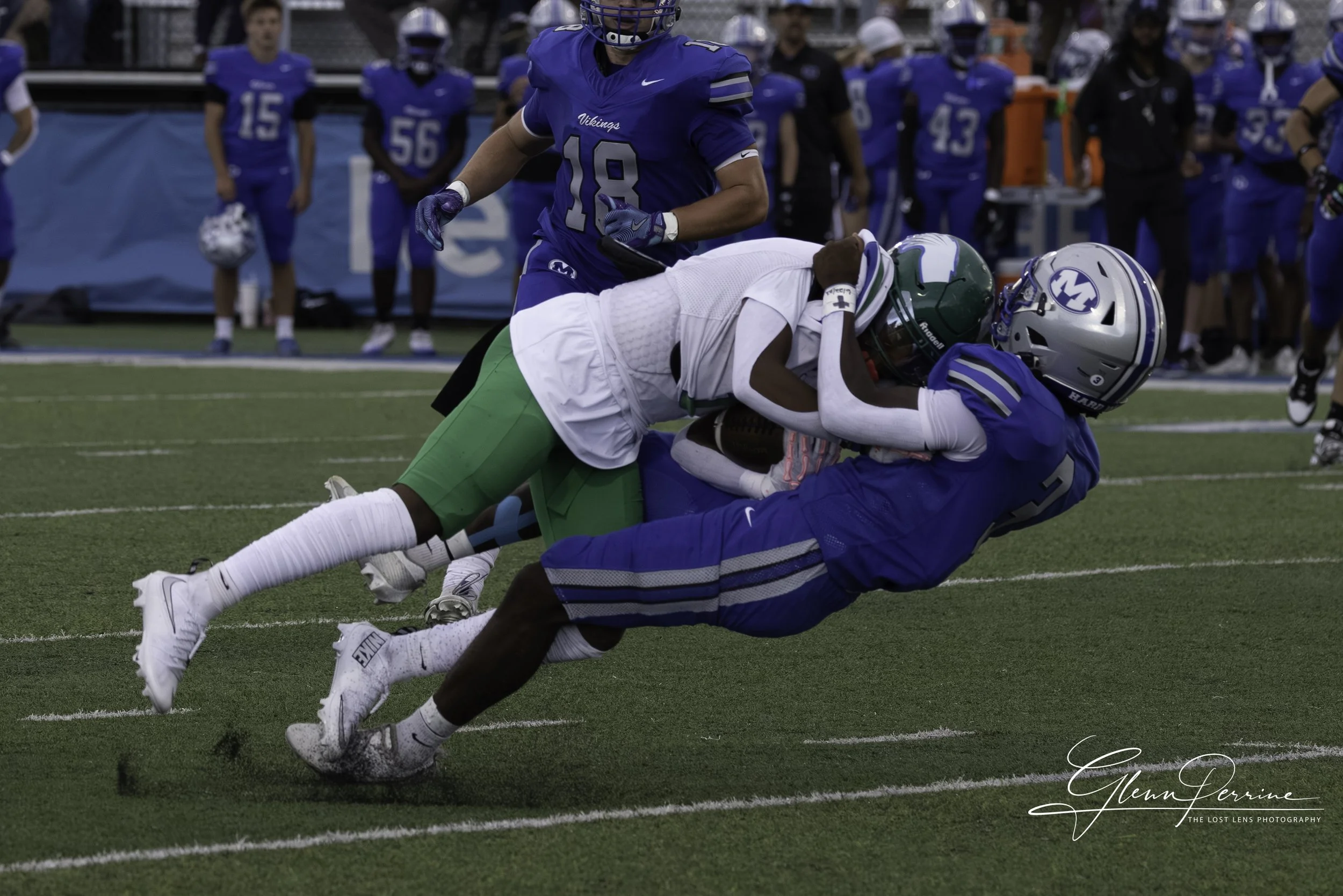 A football player in a blue uniform and a football helmet tackles a player in a green uniform who is holding a football, on a football field with other players and spectators in the background.