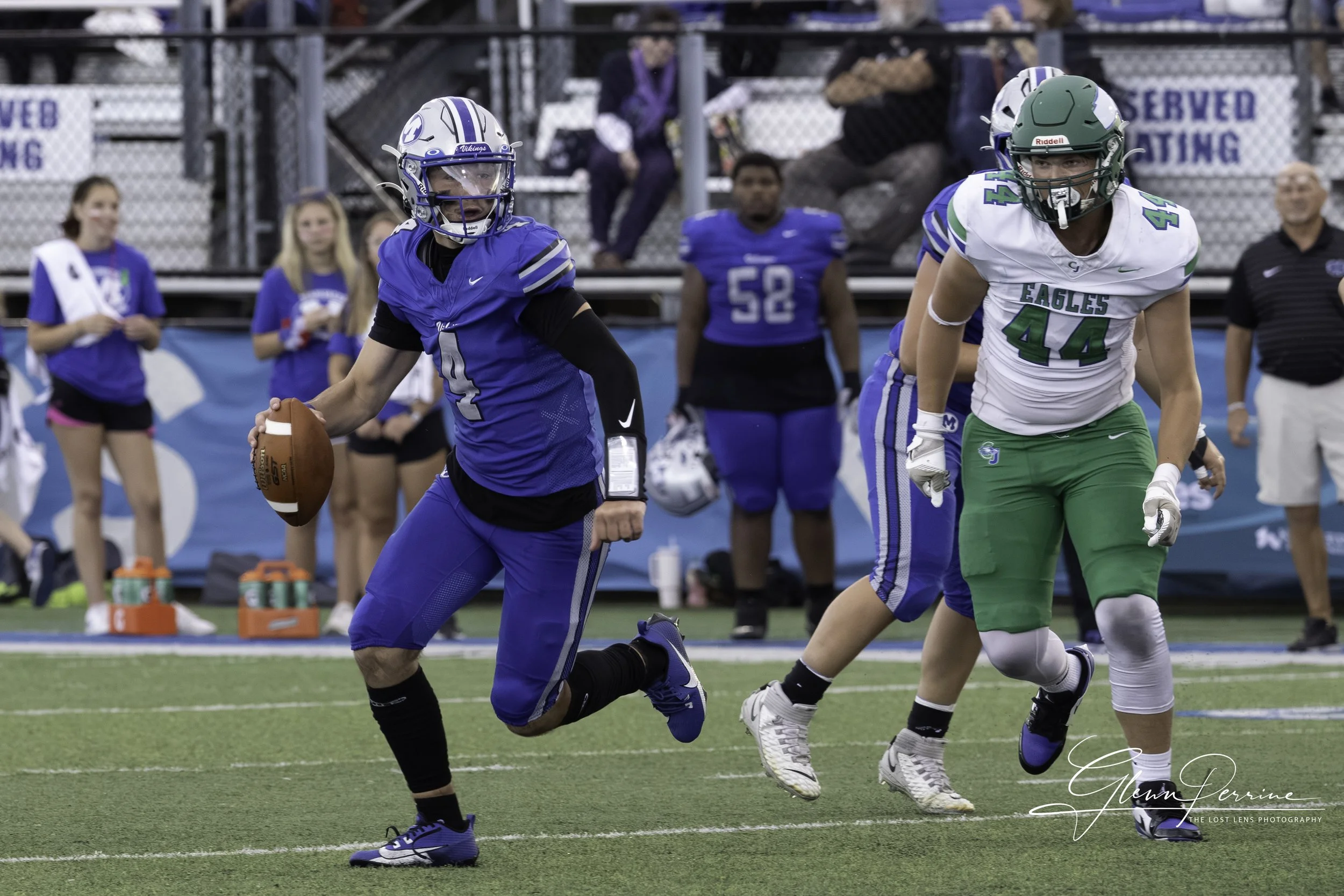 Football players on the field with a player holding the ball running while others move around him. Spectators and team members in the background.