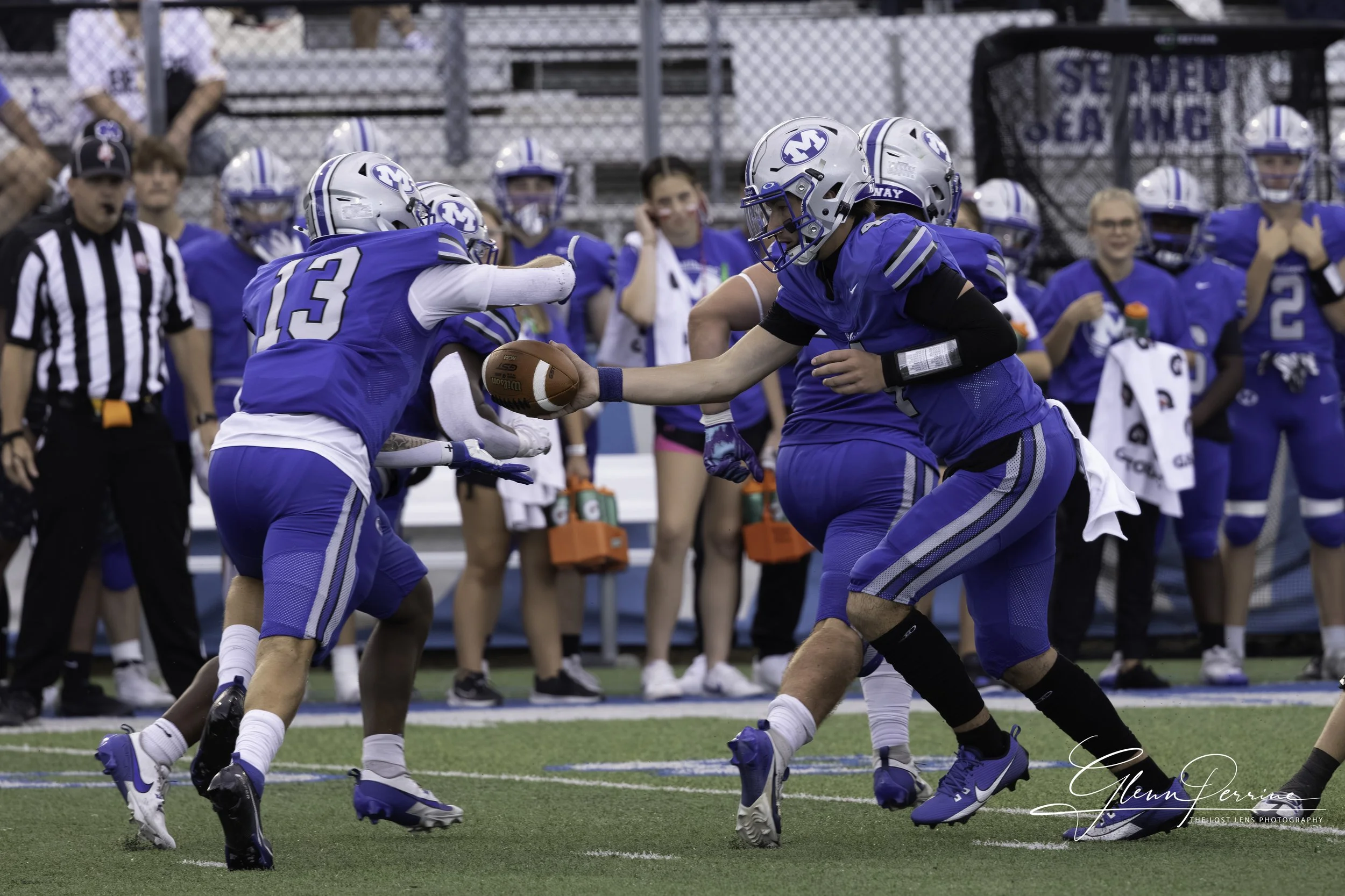 Two football players in blue jerseys and helmets engage in a tackle on the field while other players and spectators watch from the sidelines.