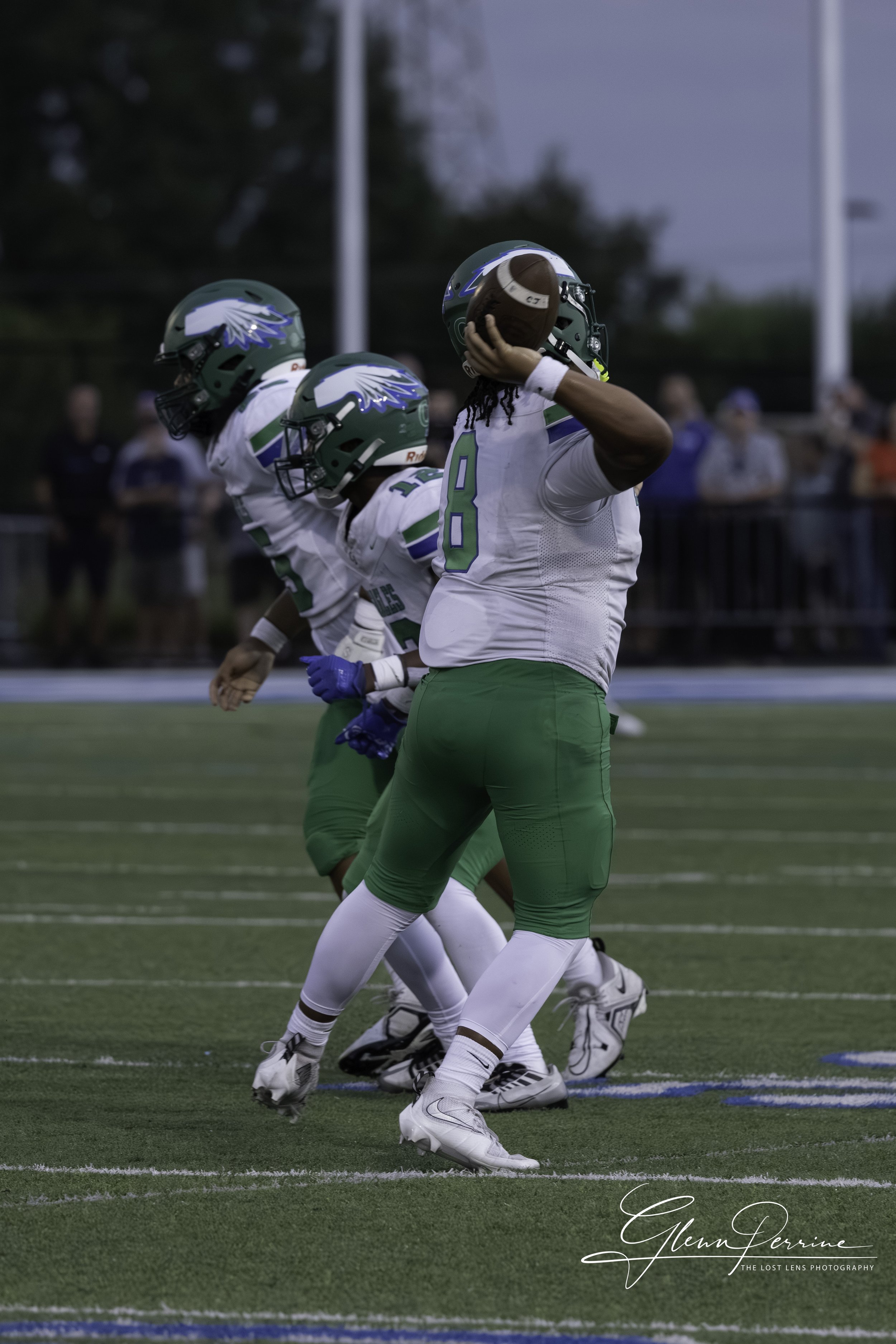 Football player in green and white uniform catching a football during a game, with two teammates in similar uniforms in the background, on a football field.
