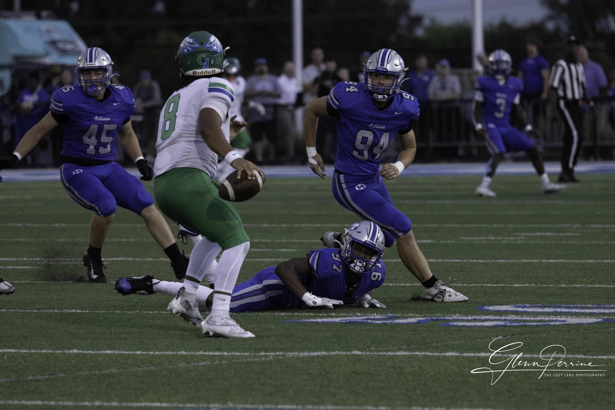 A football game in progress with players from two teams, one in blue and the other in white and green. The player in white with green pants is holding the football, trying to run. Several players in blue are surrounding him, with one player on the gr