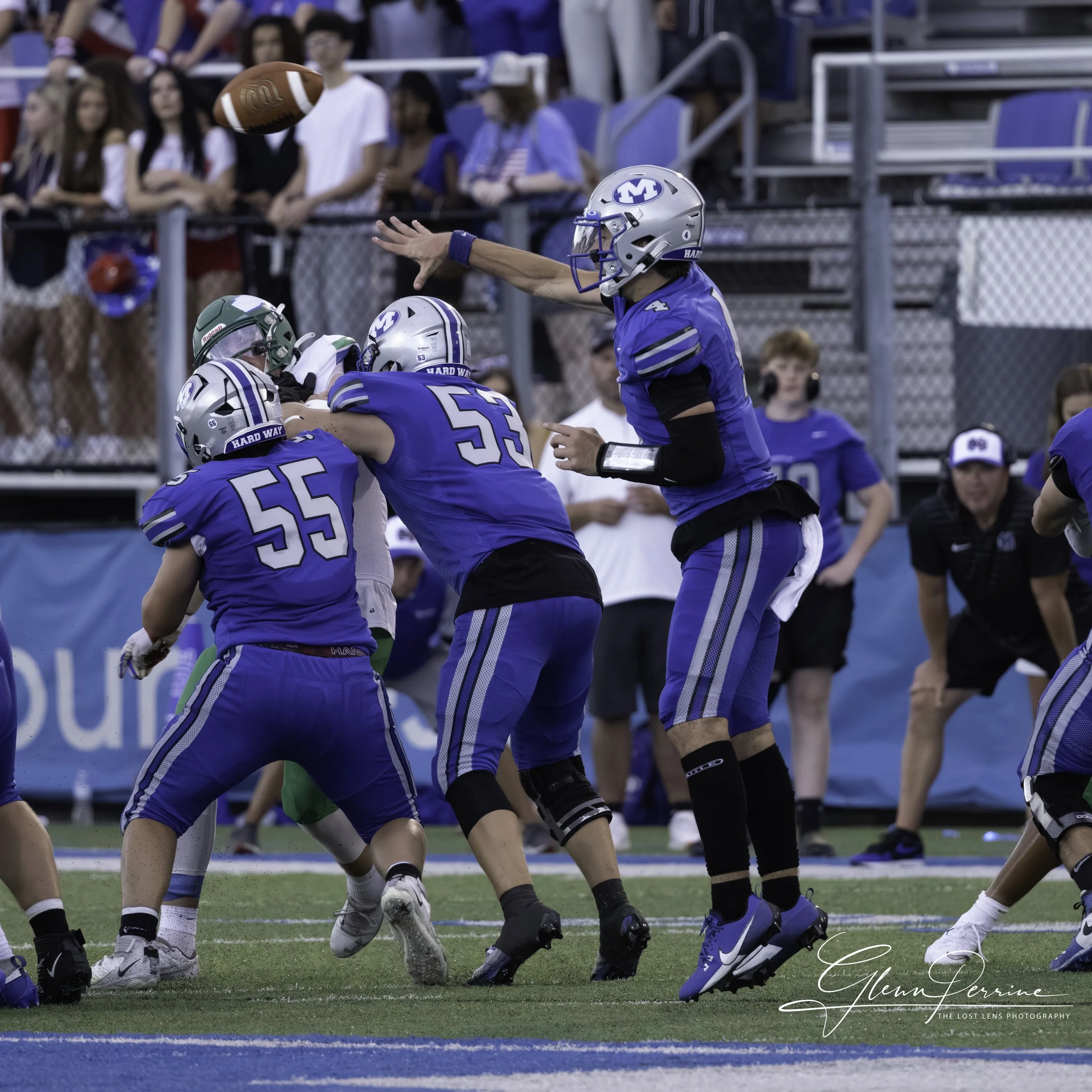 Football players in blue and green jerseys engaged in a play on the field, with spectators watching in the background.