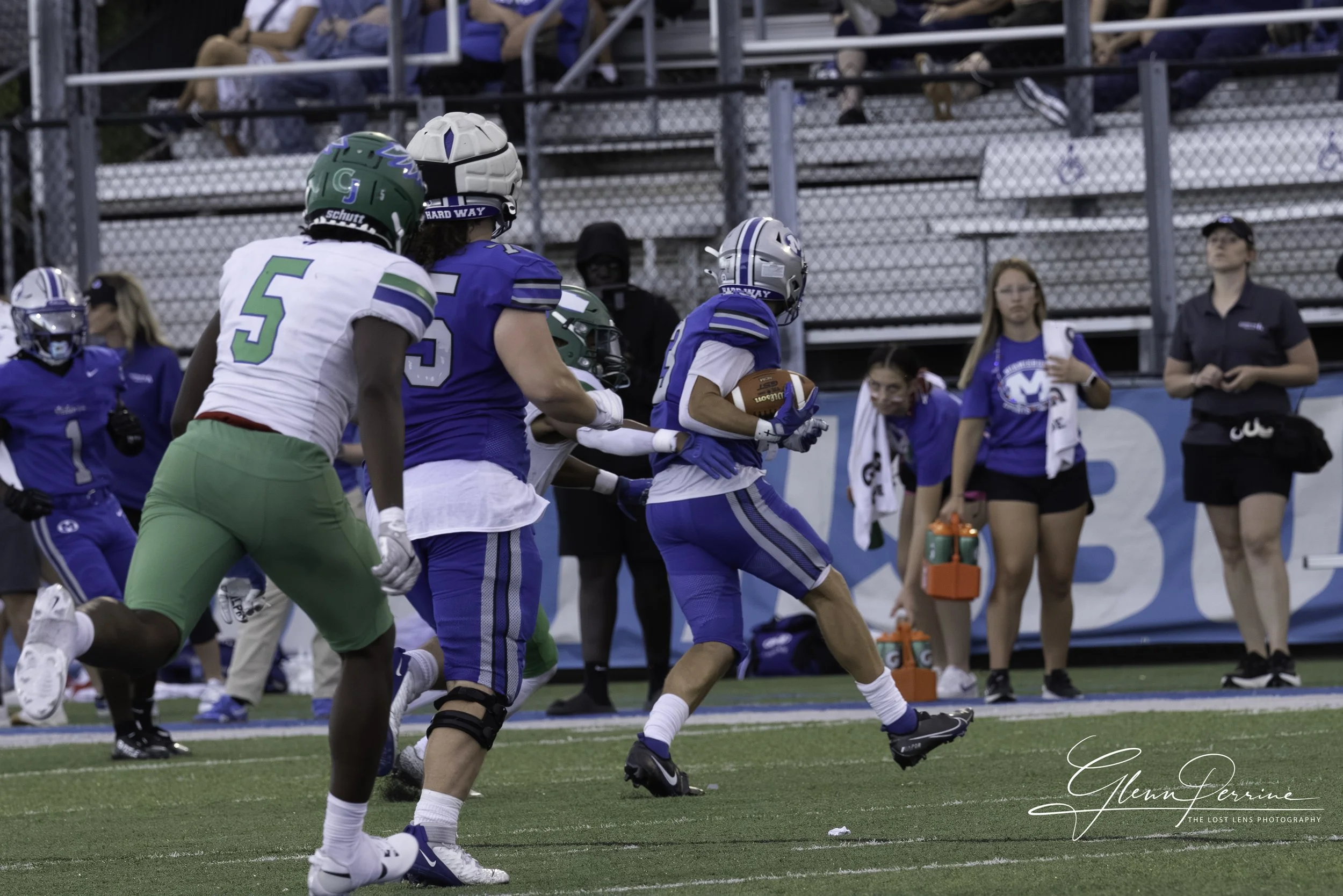 A football game with players on the field, one player in blue running with the ball, and others in white and green trying to catch him. Spectators and team staff are on the sidelines.