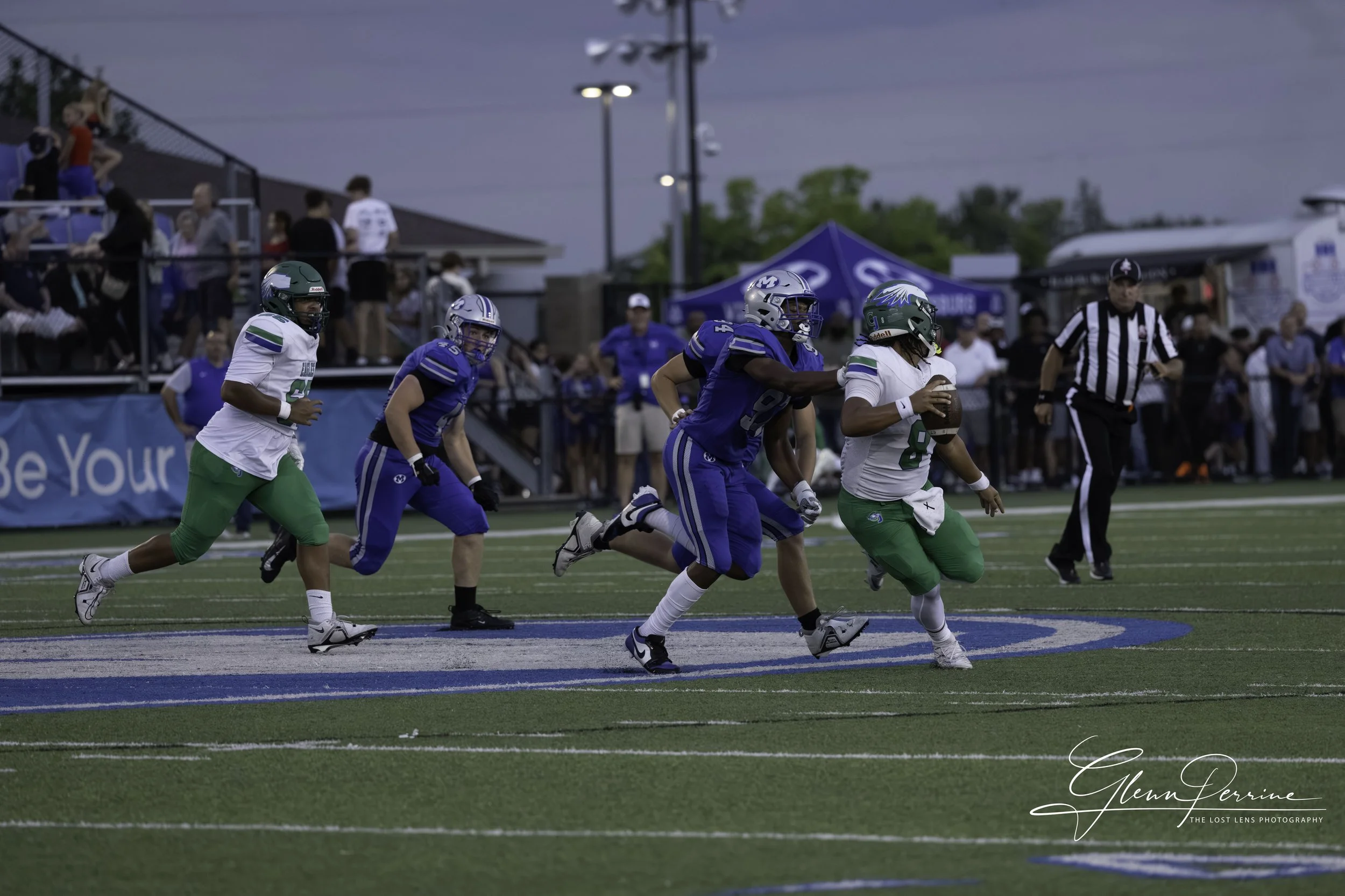 Football players during a game, with one player running with the ball as others chase behind, spectators in the stands, and a referee on the field.