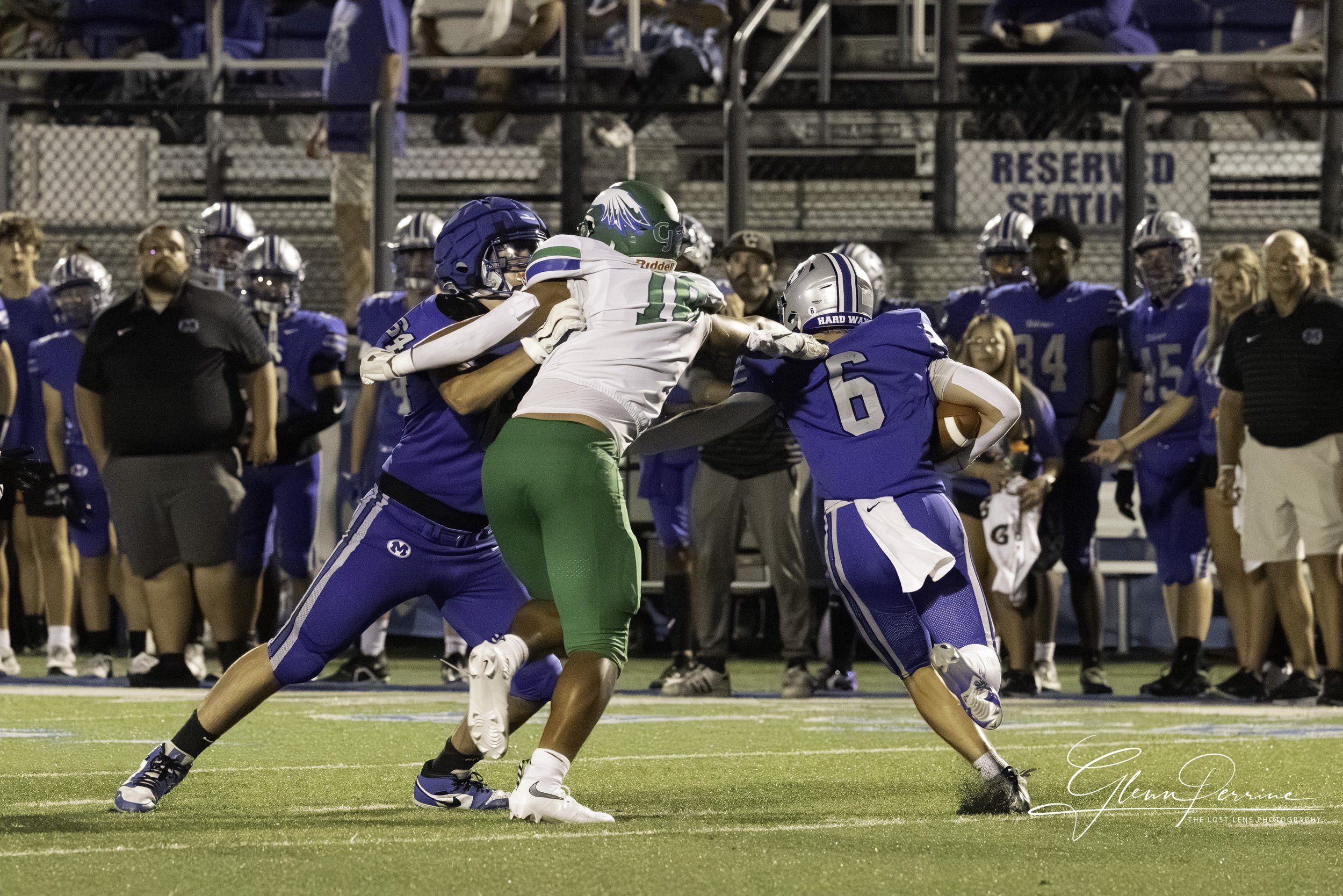 Two football players, one in white with green pants and the other in blue, tackle a player in blue holding a football during a game. Several players and coaches are visible in the background on the sideline. The game is taking place on a grassy field