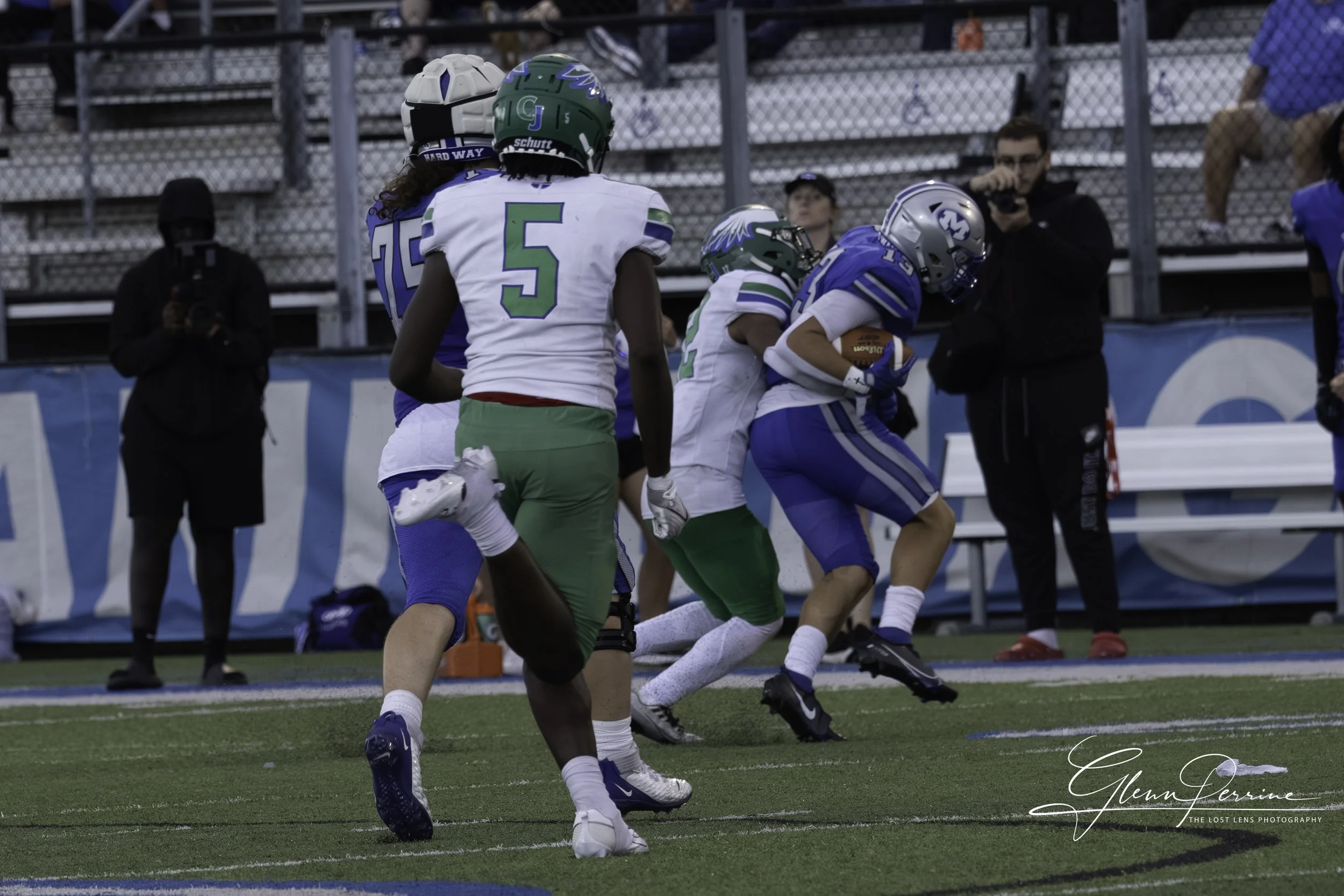 Football game with players in blue, green, and white uniforms on the field, some running and others near the sideline, with photographers and spectators in the background.