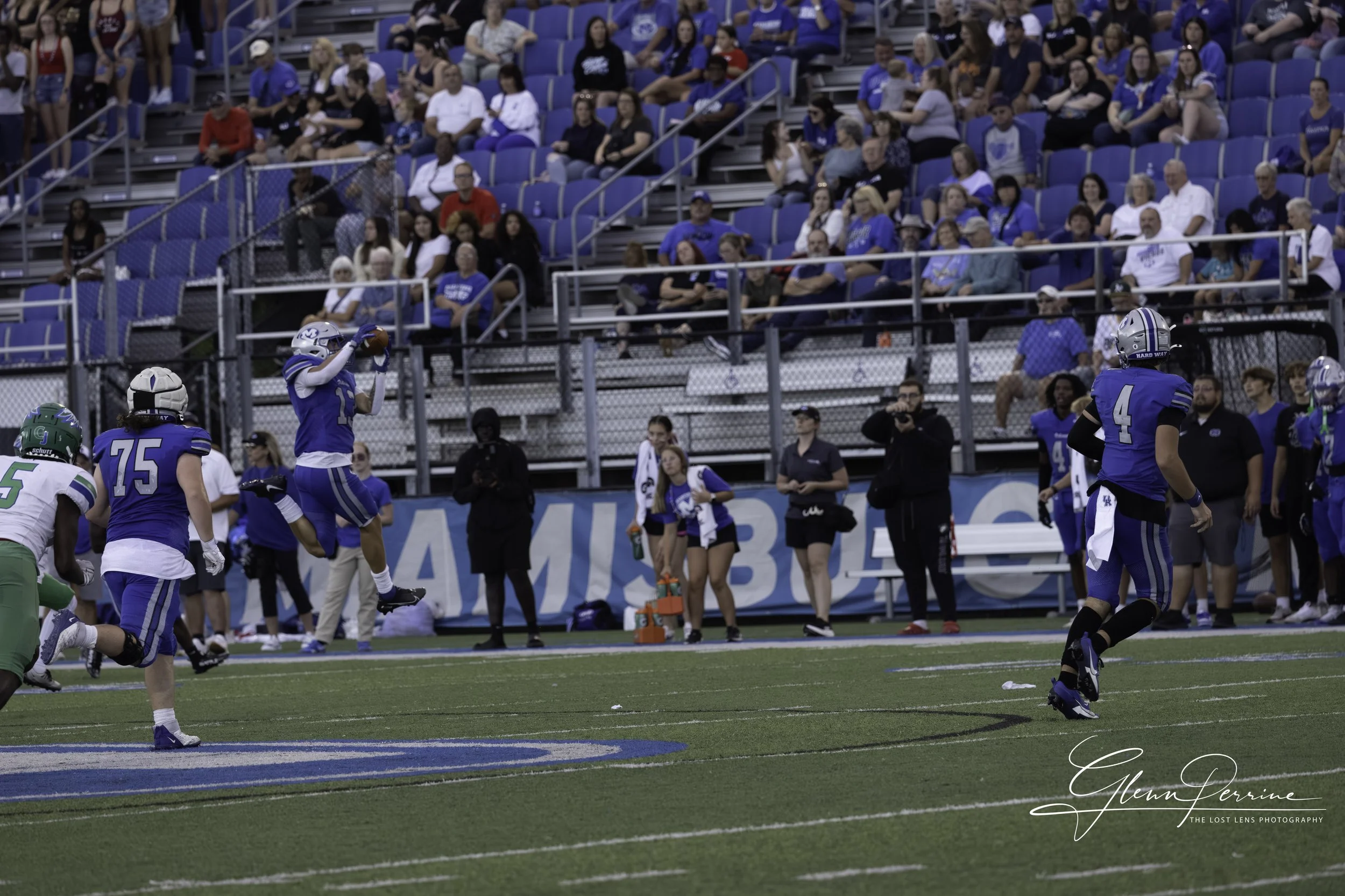 A football player in a blue jersey is catching a ball near the sideline during a game. Other players, spectators, and photographers can be seen in the background.