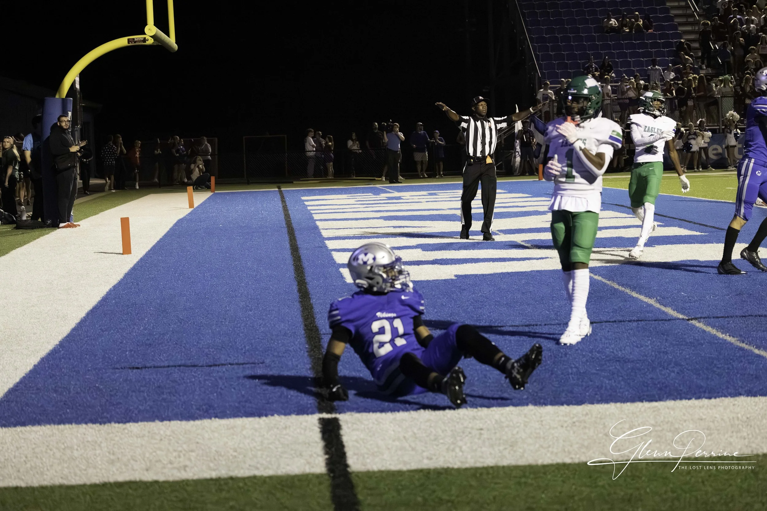 A football game scene at night with players near the end zone; a player in blue with the number 21 is sitting on the ground near the sideline, while other players in white and green are celebrating or celebrating touchdowns; a referee with arms raise