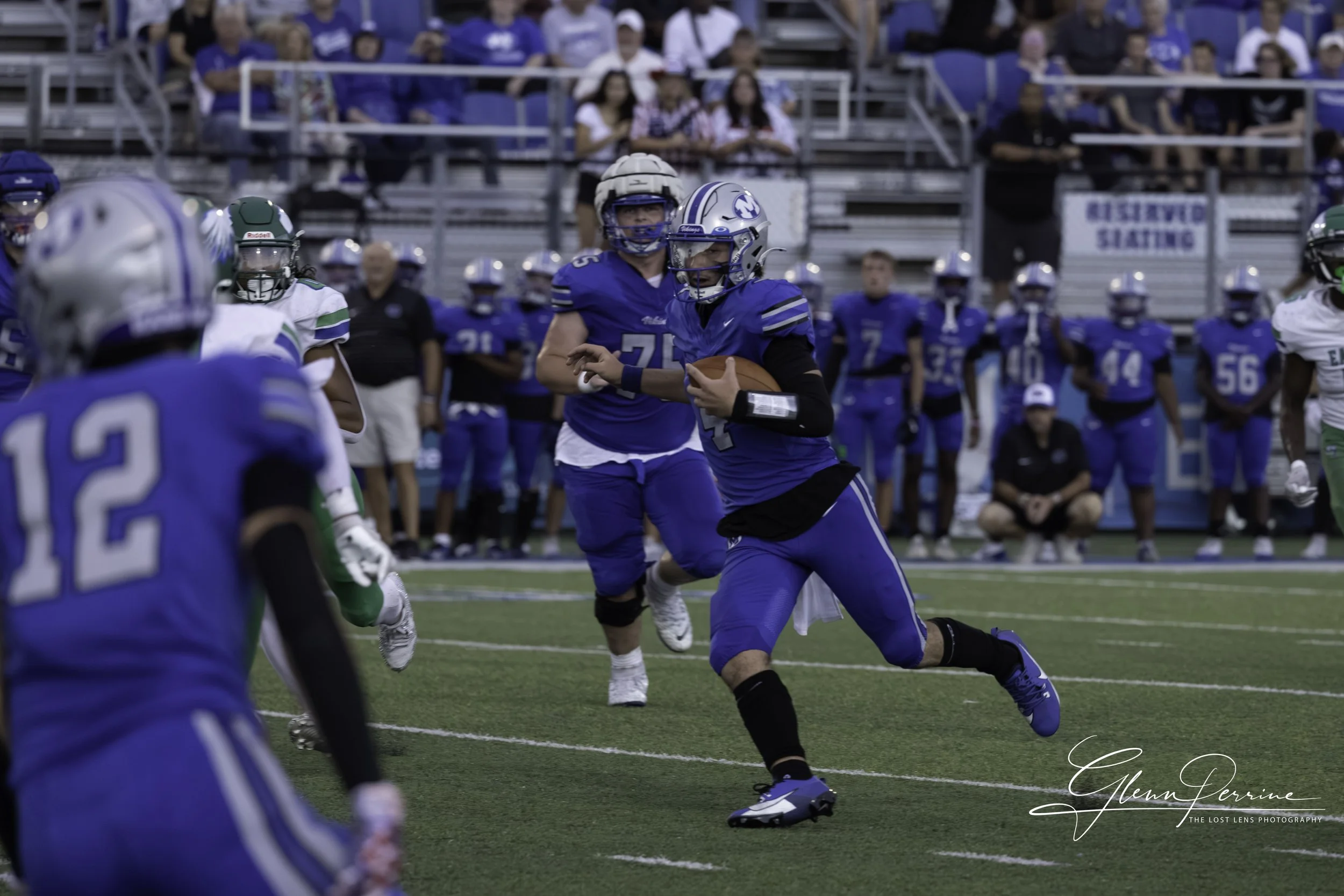 A football game in progress with players in blue and white uniforms. A player in blue with the number 4 is running with the ball, while other players, including someone with the number 12, are positioned nearby. Fans are in the stands in the backgrou
