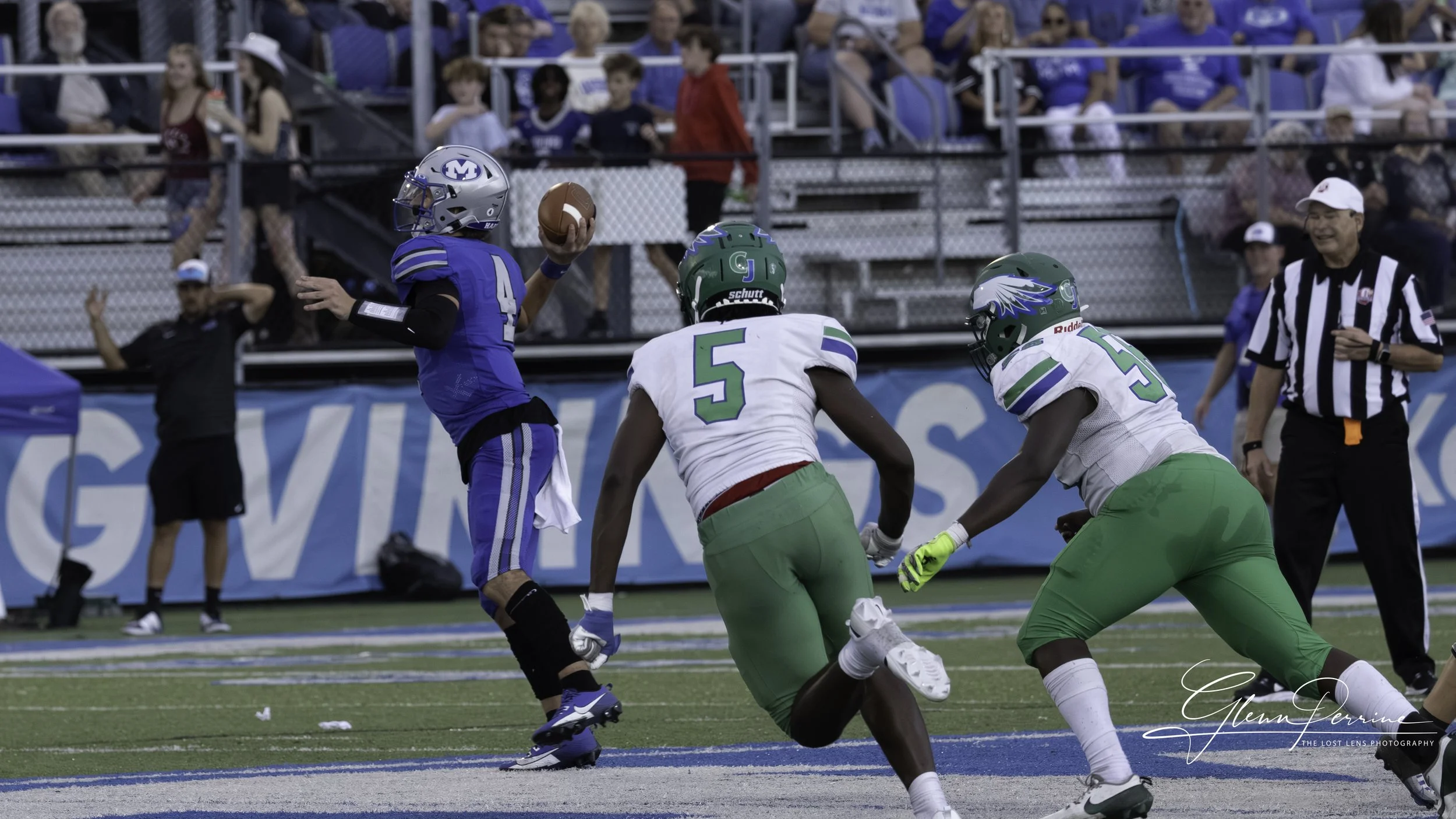 A football player in a blue uniform with the number 4 is throwing a football during a game, surrounded by two players in white and green uniforms, and an official in a black and white striped shirt. The background shows spectators in the stands.