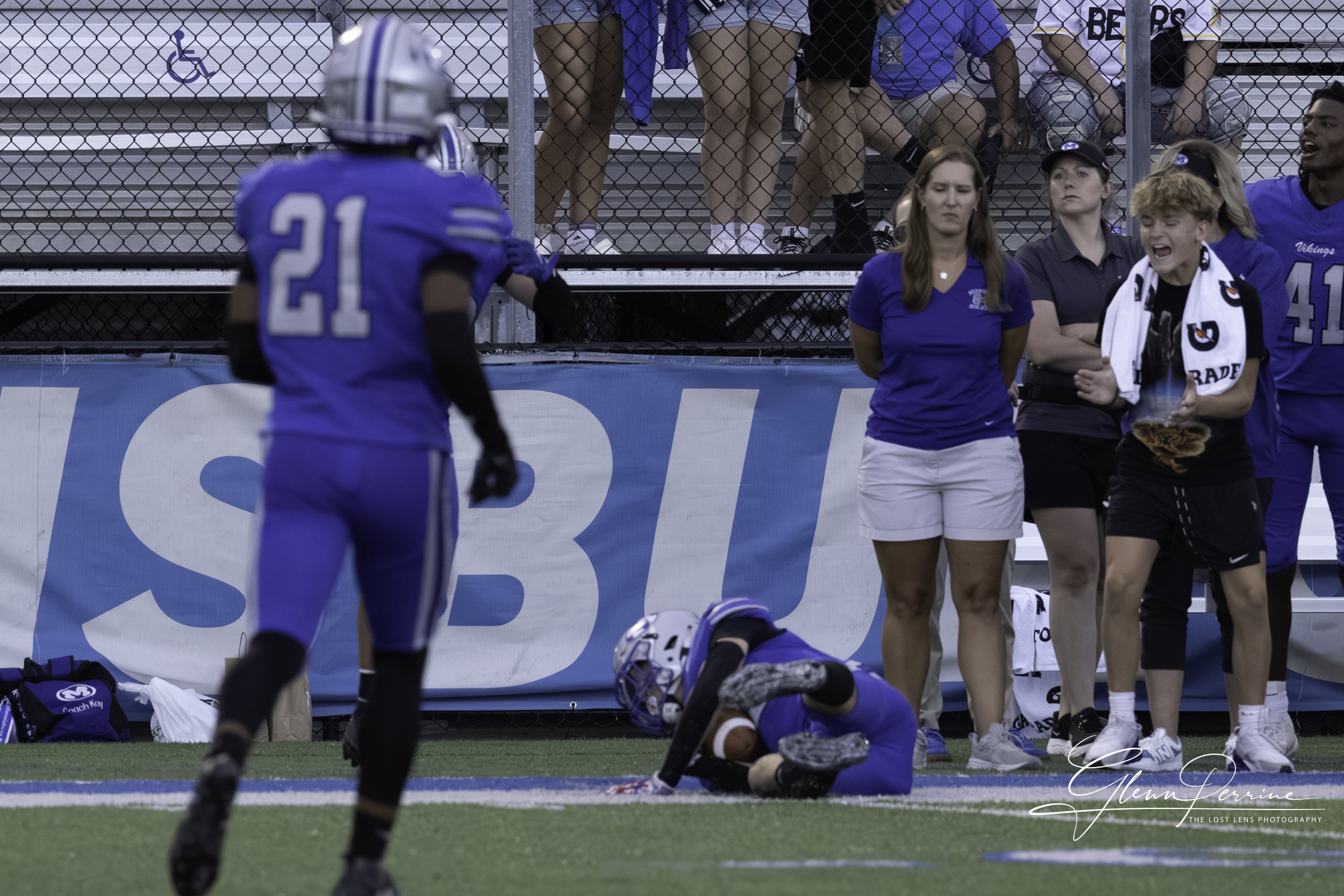 A football player in a blue uniform lying on the ground holding a football near the end zone, with a teammate in a blue jersey and a black and white scarf with a basketball logo watching nearby, and other people and spectators behind a fence.