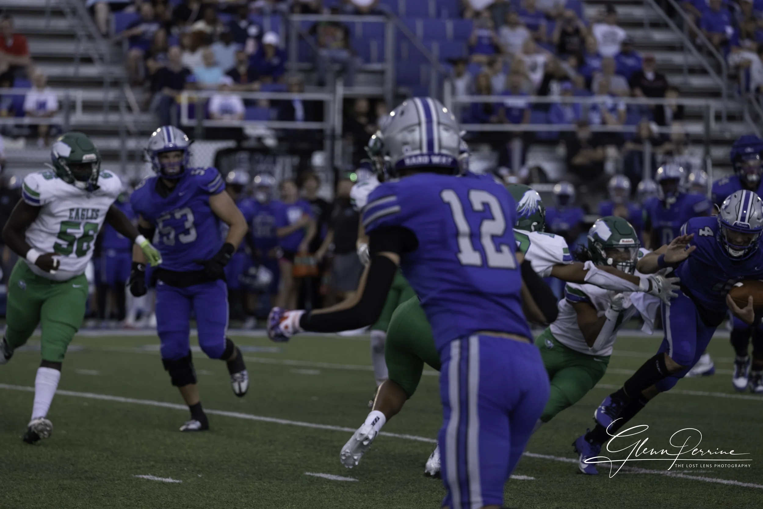 An American football game with players in blue and green uniforms on the field, some running and others tackling, with spectators in the stands in the background.
