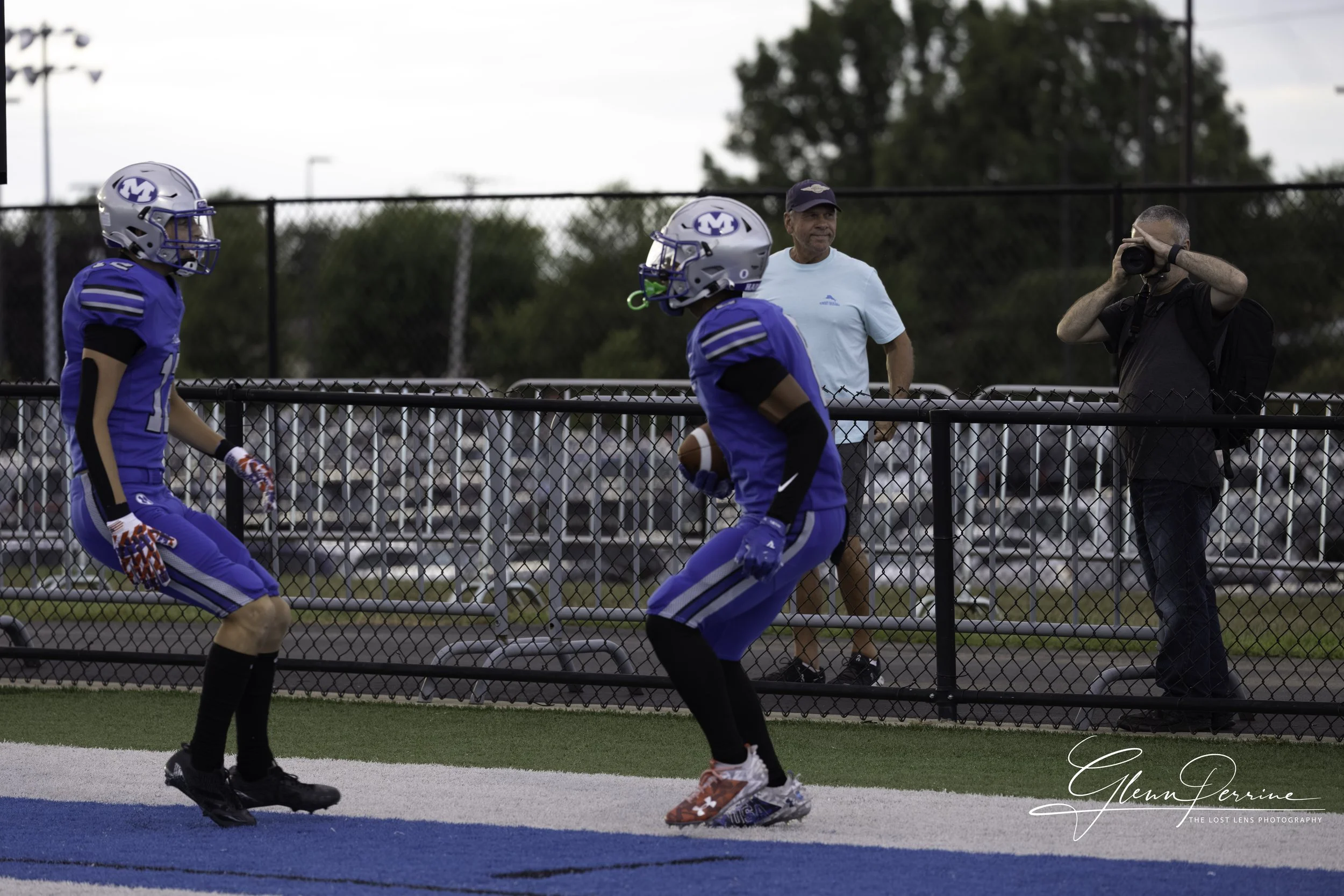 Two football players in blue uniforms and helmets on the field, one holding a football, with a coach, another person watching with a camera, and a fenced sports field background.