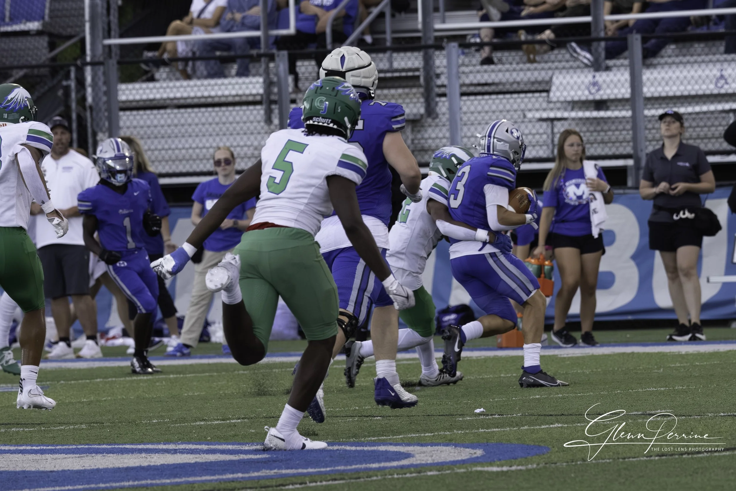 American football game with players running on the field, some wearing white and green uniforms and others in blue, with spectators and coaches in the background.