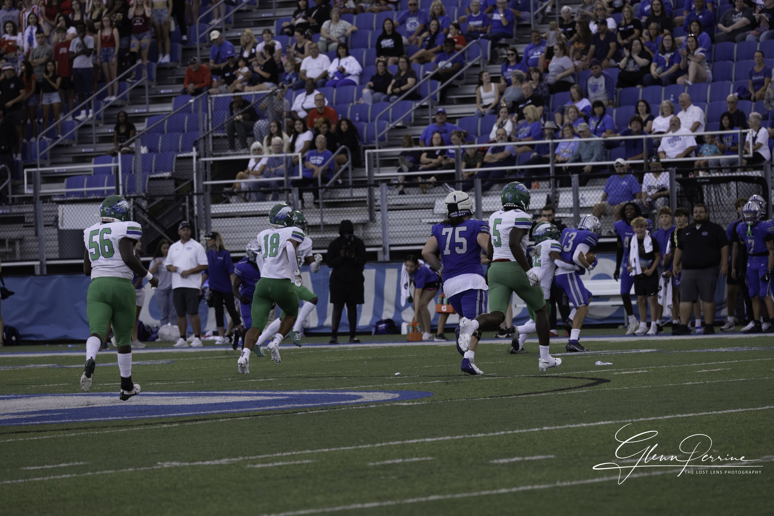 Football players in white and green uniforms running on the field during a game, with spectators in the stands.