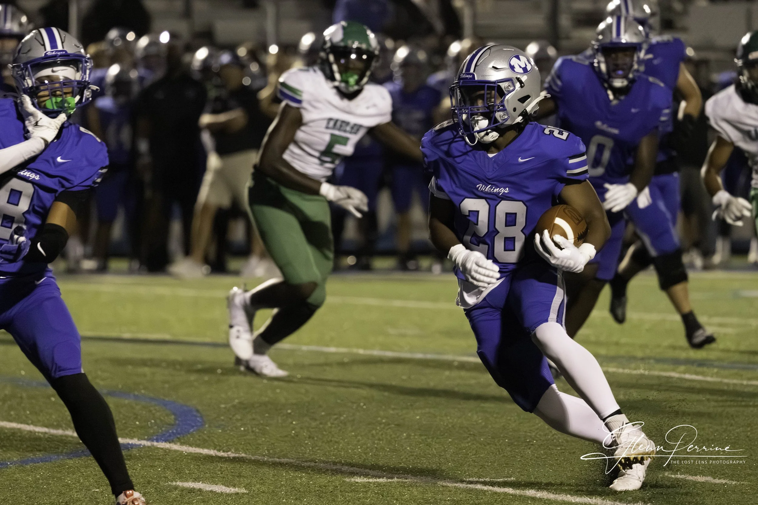A football player in a blue uniform with the number 28 is running with the ball. Other players and spectators are visible in the background.