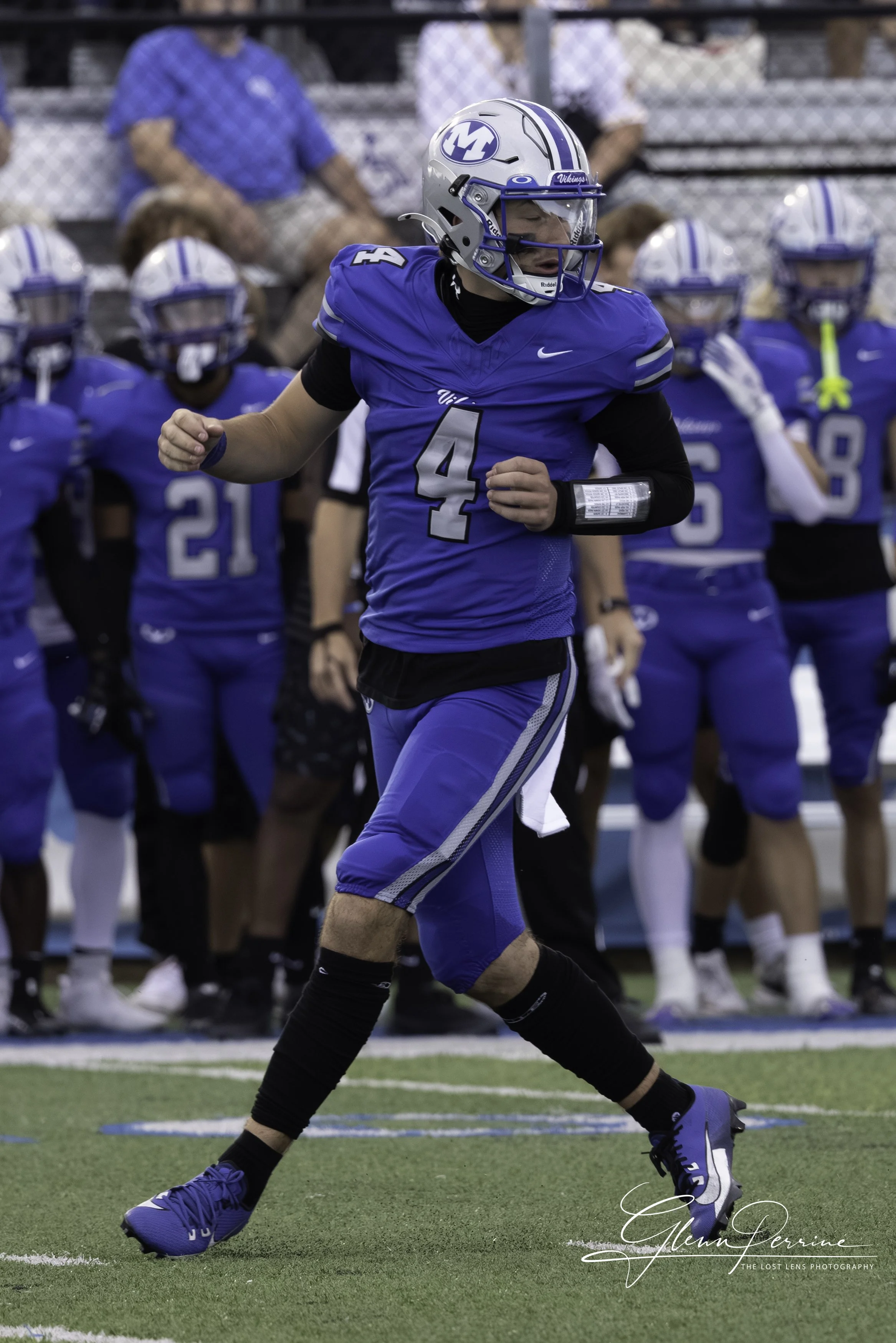 A football player in a blue uniform with number 4 on it, running on the field during a game, with teammates and spectators in the background.