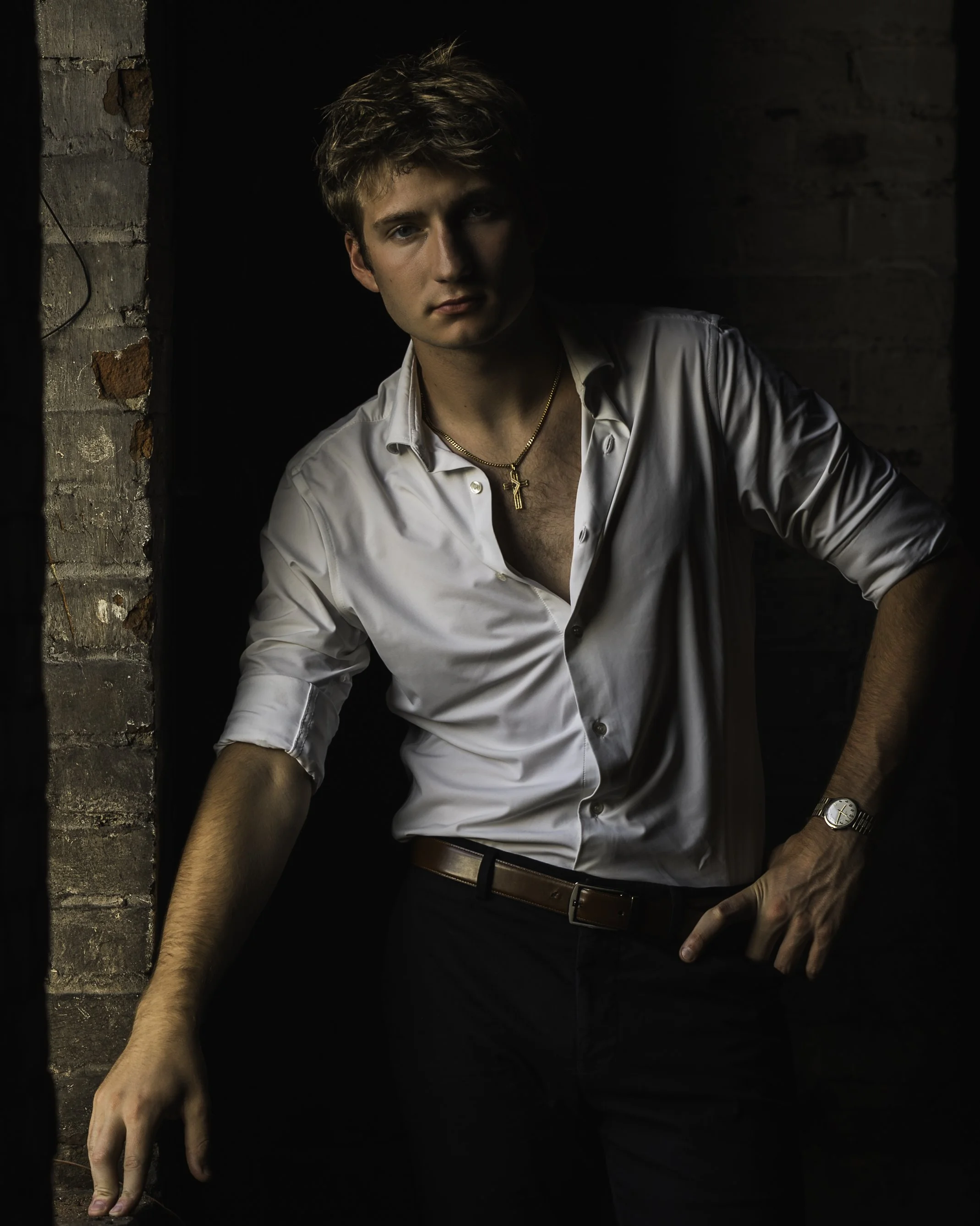 A young man with tousled hair wearing a white button-up shirt, gold necklace, and wristwatch posing against a dark background near a brick wall.