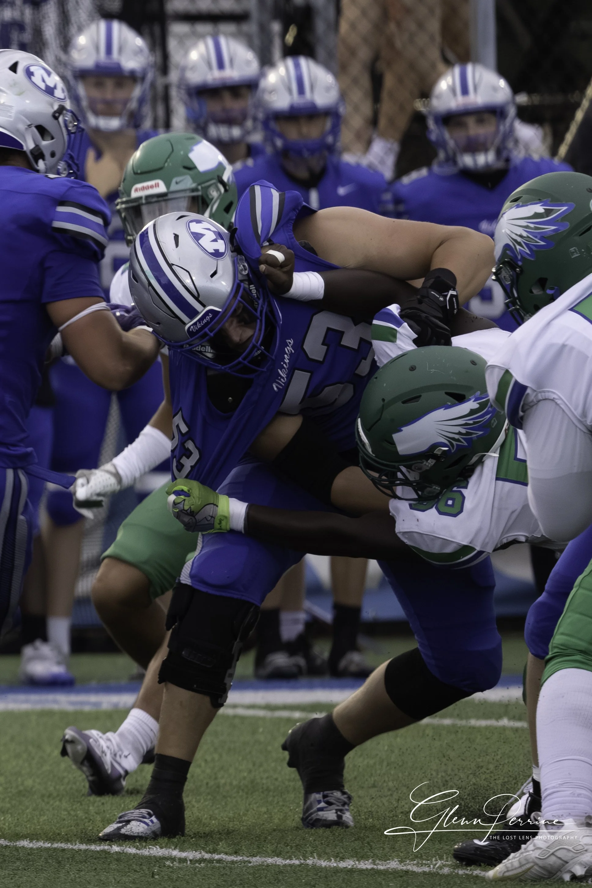 An American football game with players in blue and white uniforms engaging in a tackle, with some players in green and white uniforms attempting to block or defend.