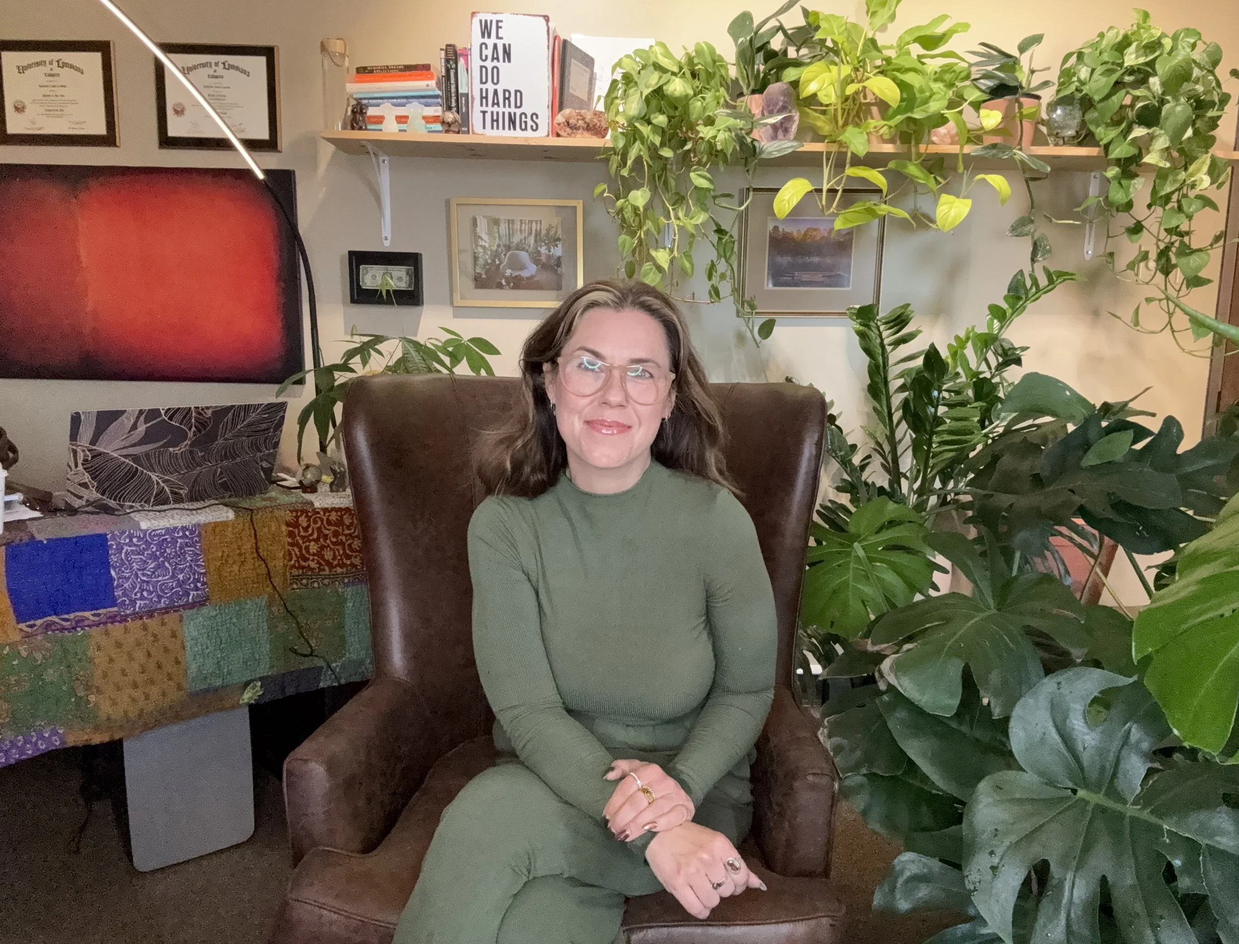 Kate Lemoine, LPC sitting in a leather armchair in a room filled with green houseplants, framed pictures, and shelves with books and therapeutic items.