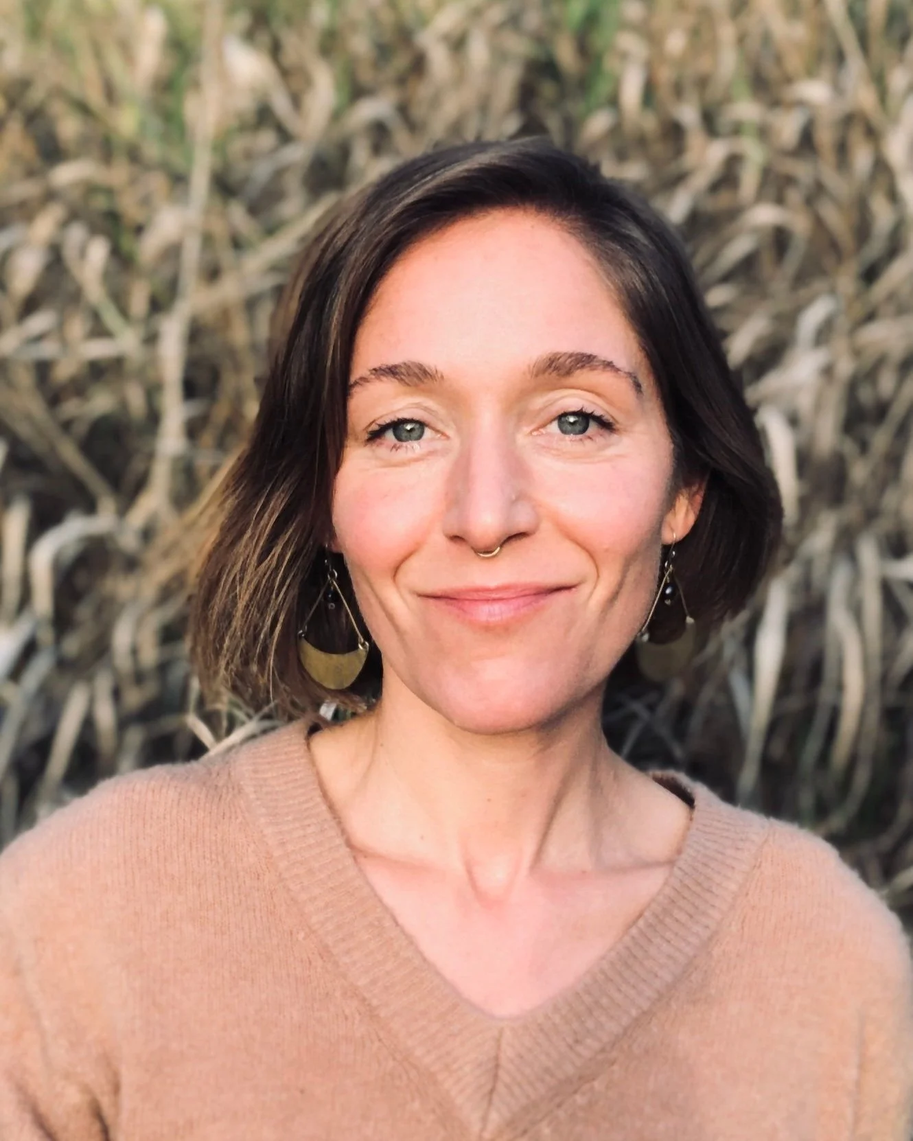 A woman with medium-length dark hair, blue eyes, and a septum piercing, wearing earrings and a beige sweater, smiling outdoors with a background of dried reeds or grass.