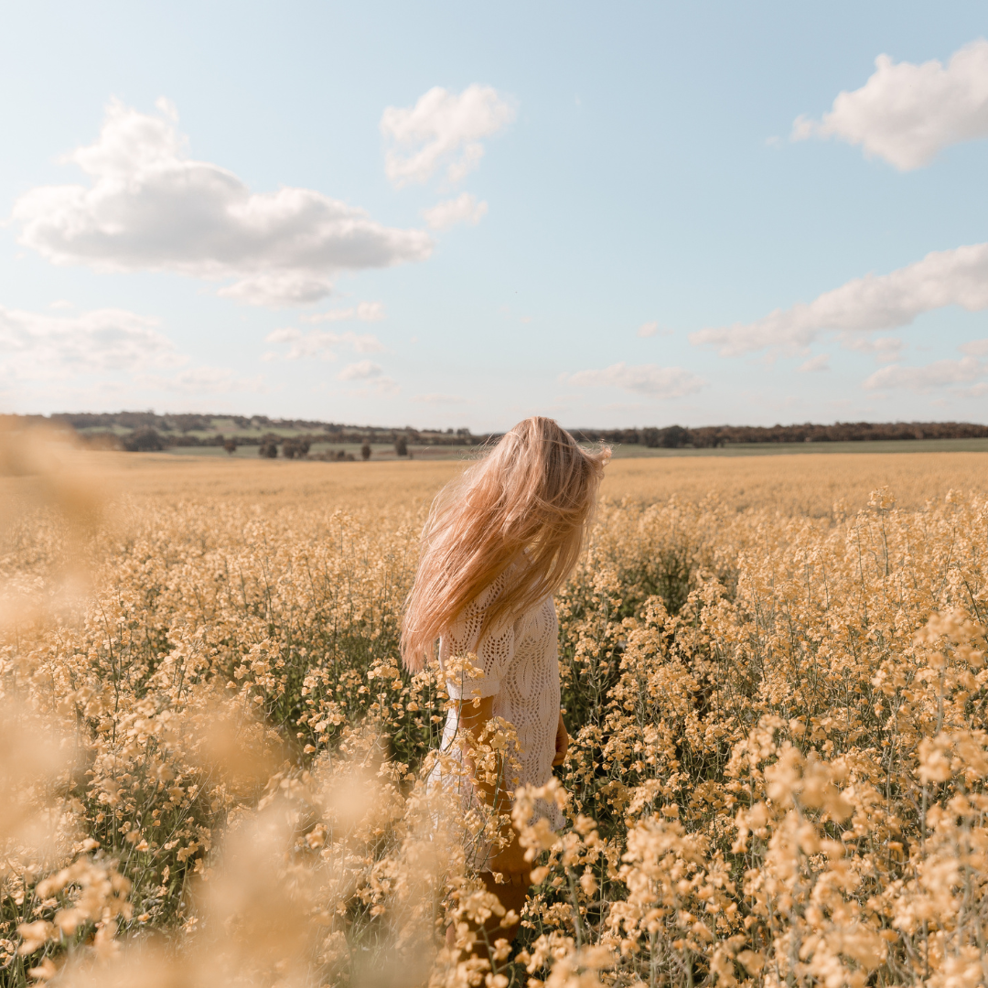 A woman with long blonde hair wearing a white sweater standing in a vast field of yellow flowers under a partly cloudy sky.