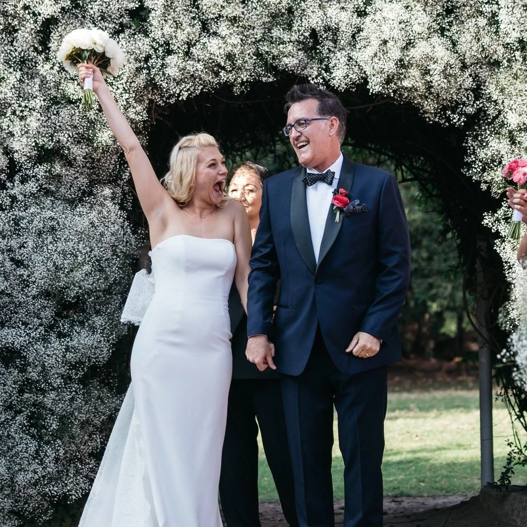 A bride in a white wedding dress celebrating with her groom in a tuxedo under a floral arch, holding a bouquet in her raised hand at an outdoor wedding ceremony.