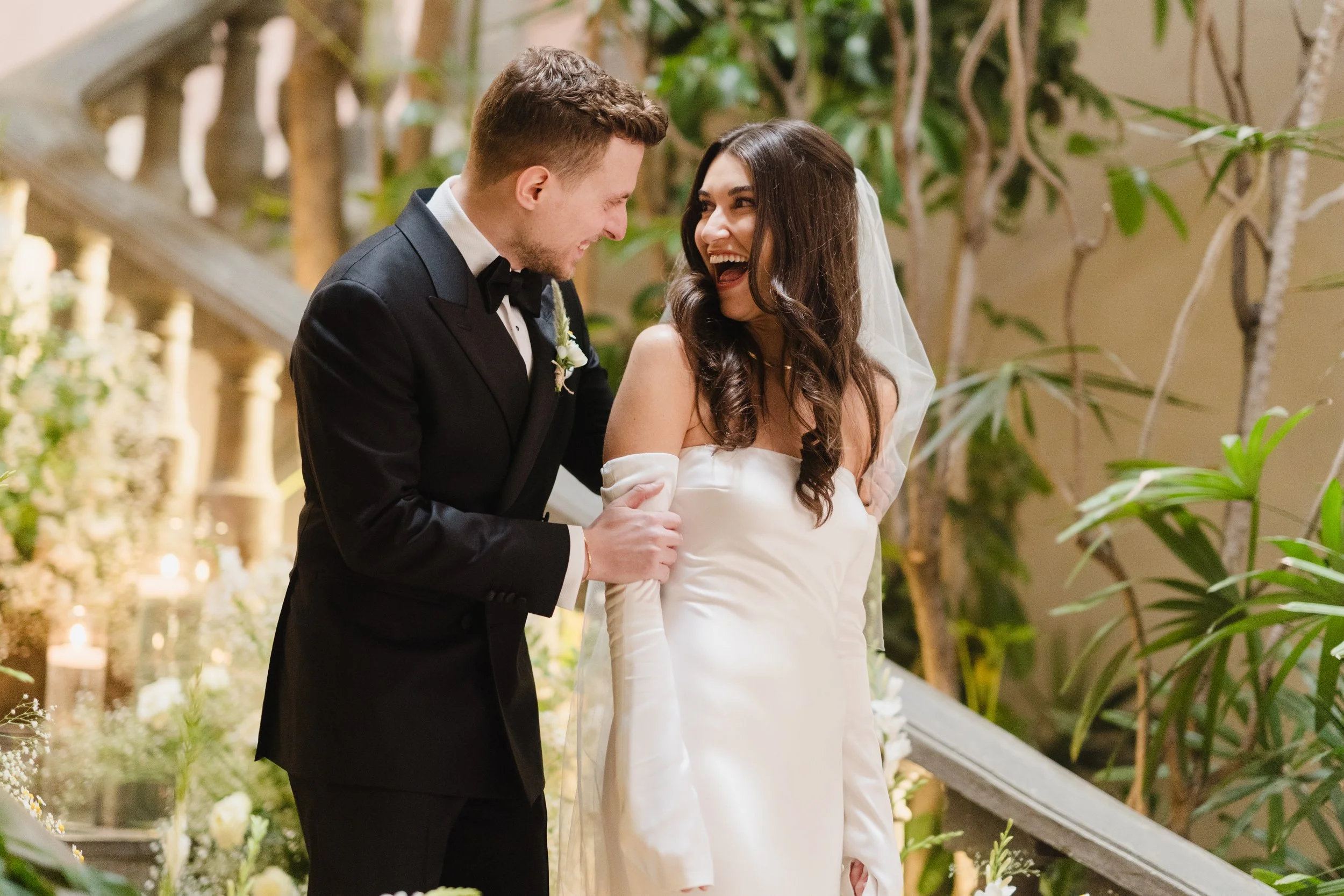 A bride and groom share a joyful moment on their wedding day, standing on a staircase surrounded by lush greenery and flowers.
