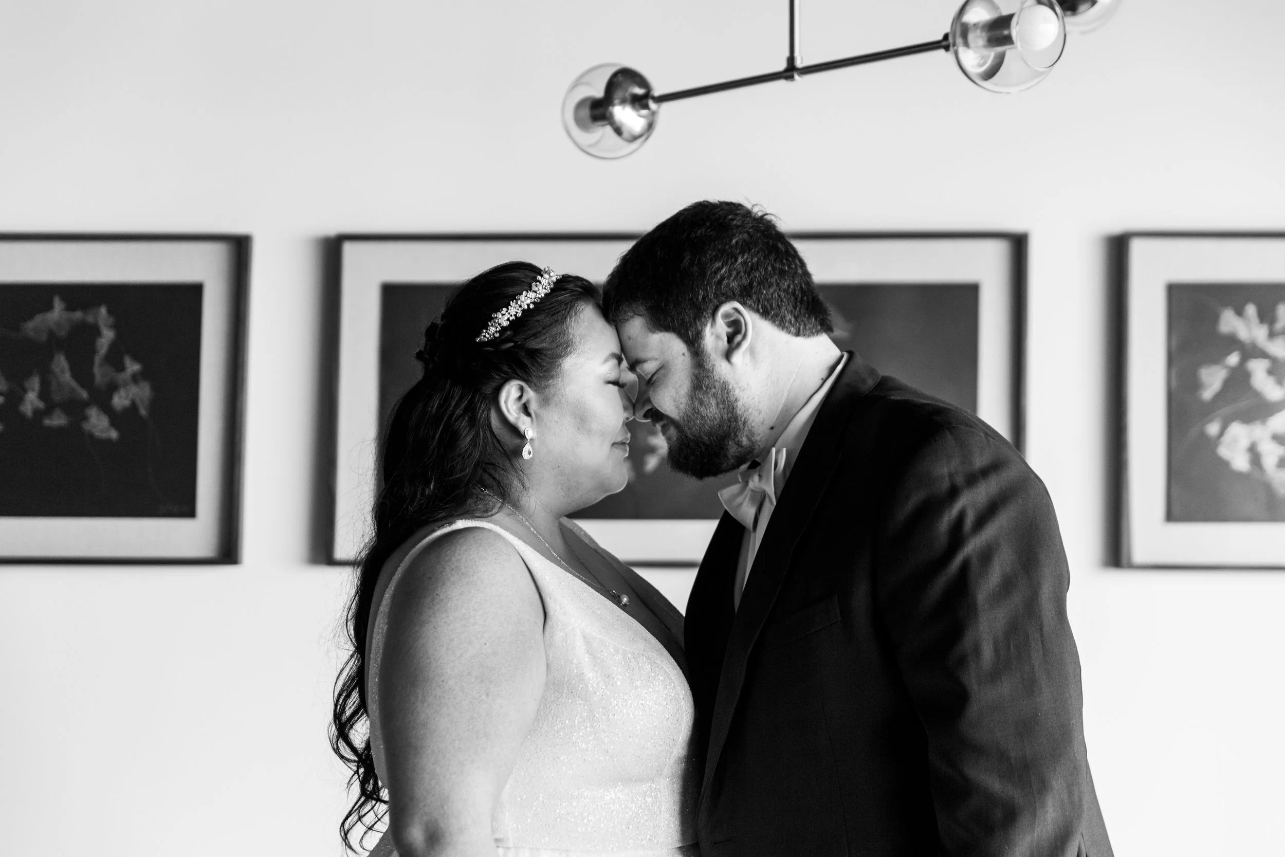 A black-and-white photo of a couple, a bride and groom, touching foreheads and smiling with eyes closed in an intimate moment. The background features framed artwork and modern light fixtures.