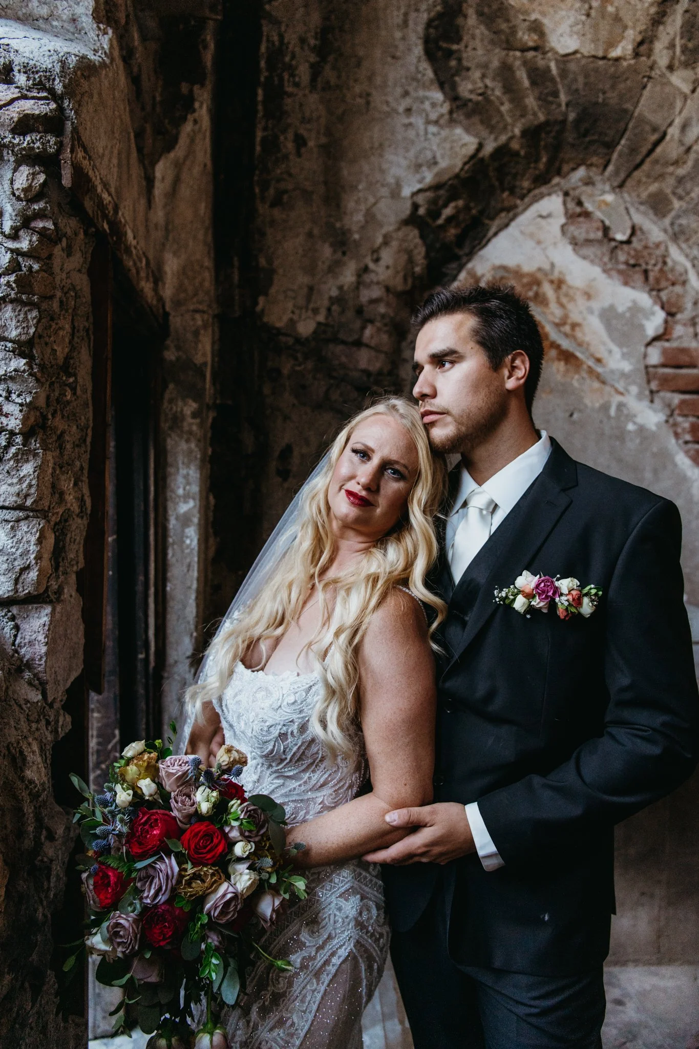 A bride and groom standing close together, the bride holding a large bouquet of red, purple, and white flowers, the groom in a black suit with a boutonnière, inside a rustic brick and stone building.