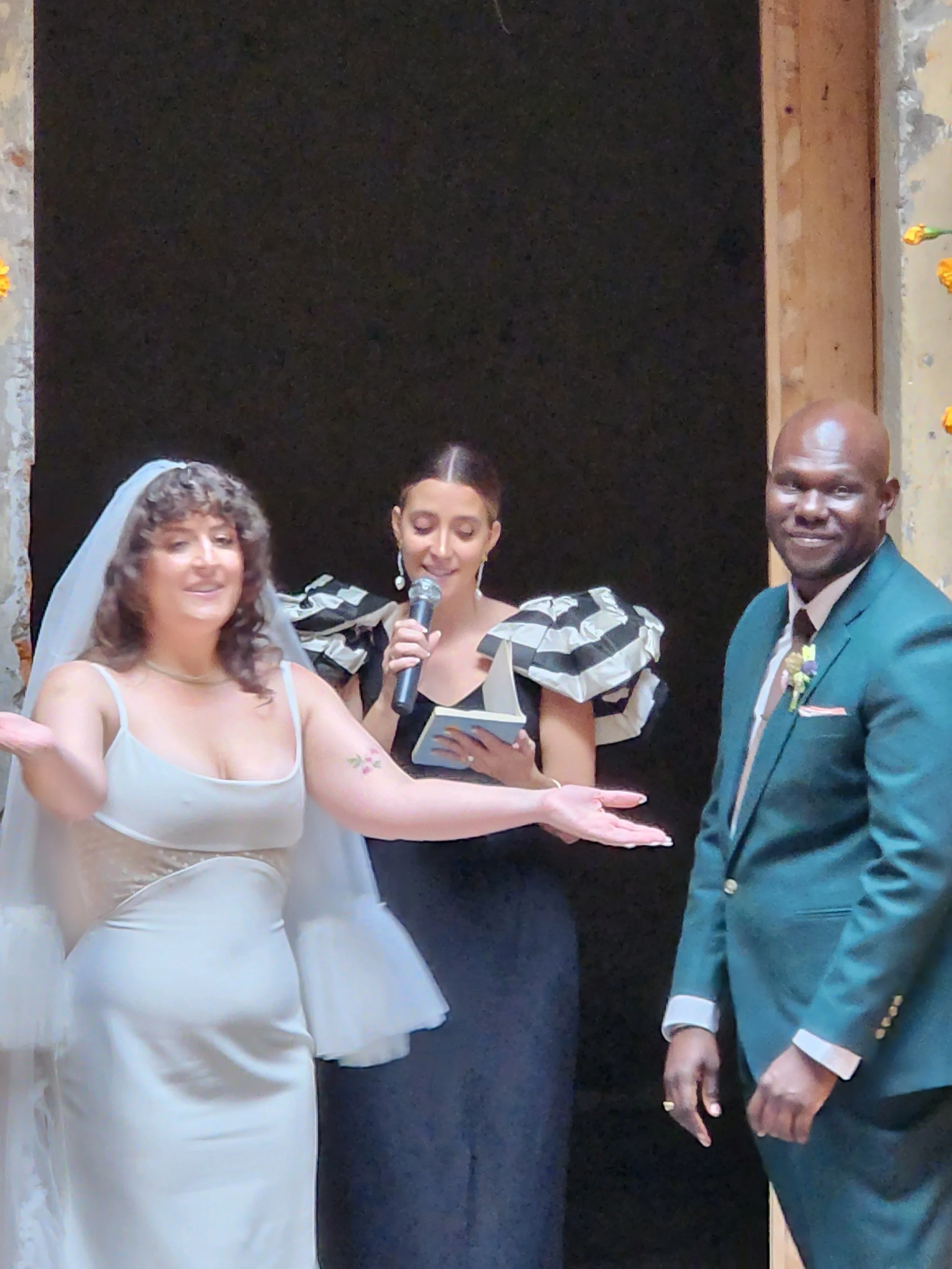 A bride in a white wedding dress with a veil, standing with a groom in a green suit, during a wedding ceremony. A woman in a black dress with puffed sleeves is holding a microphone and reading from a paper.