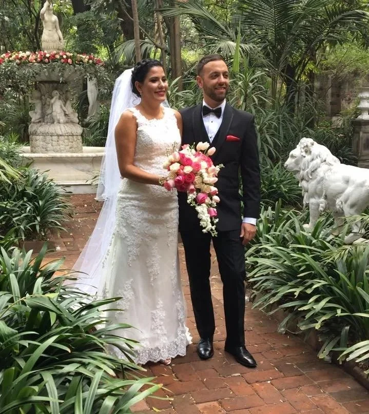 Happy bride and groom standing together outdoors in a lush garden, with the bride holding a cascading bouquet of pink and white flowers. The bride is wearing a lace wedding gown with a veil, and the groom is in a black tuxedo. There are decorative statues, including a statue of a lion and a fountain with cherubs, surrounded by green plants and trees.
