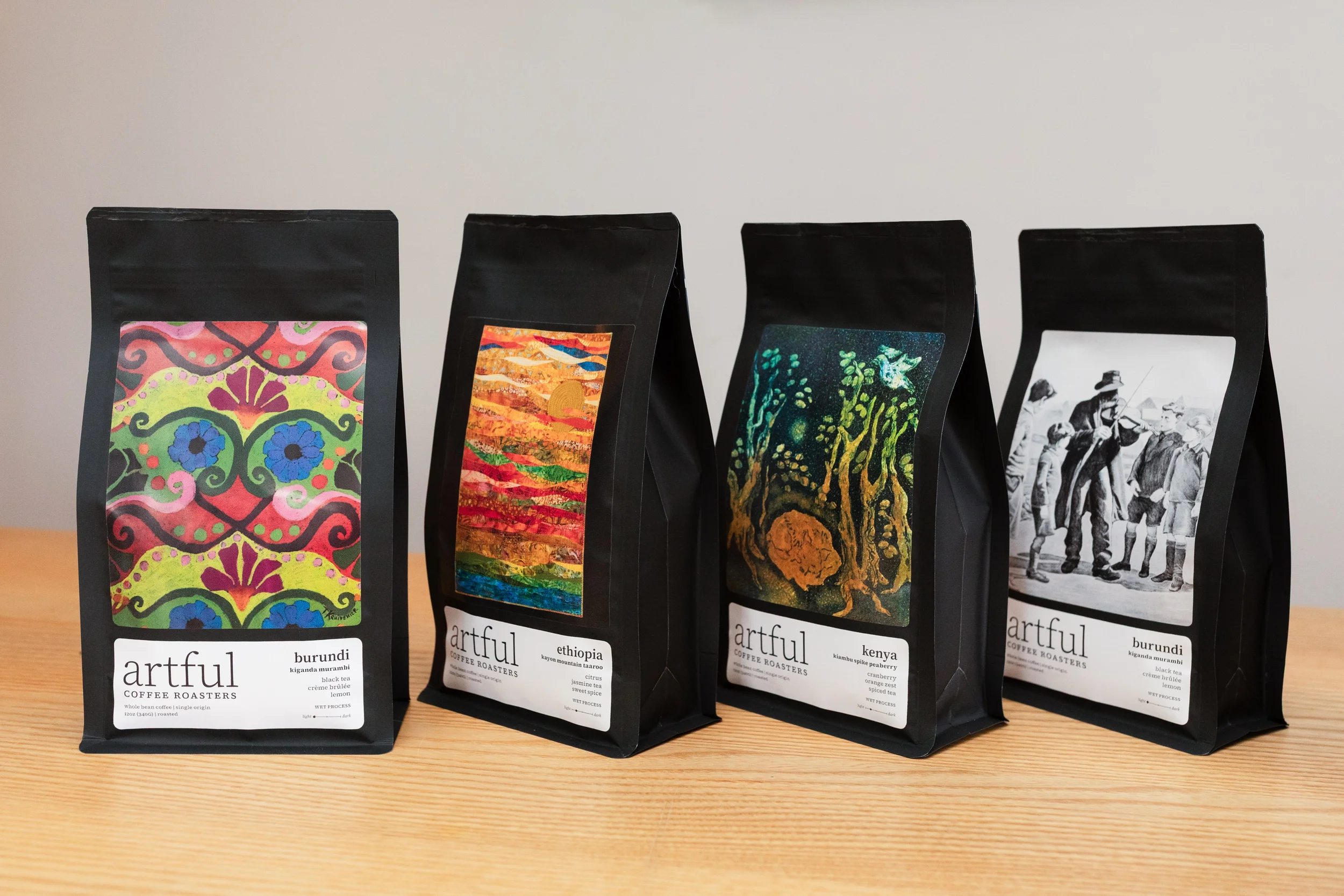 Four black coffee bags with colorful artistic labels, labeled from left to right as Burundi, Ethiopia, Kenya, and Burundi, placed on a wooden surface.